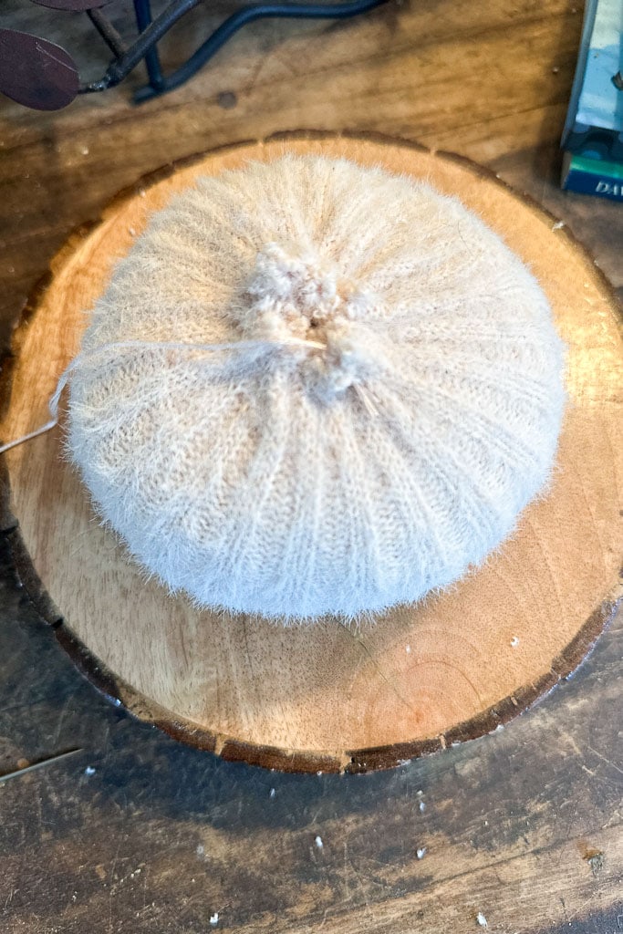 A round, soft, beige knitted sweater sleeve turns in to a sweater pumpkin sits on a wooden surface. It has a gathered top, creating a tufted appearance akin to sweater pumpkins, and is placed on a slice of a tree trunk, suggesting a natural decor setting.