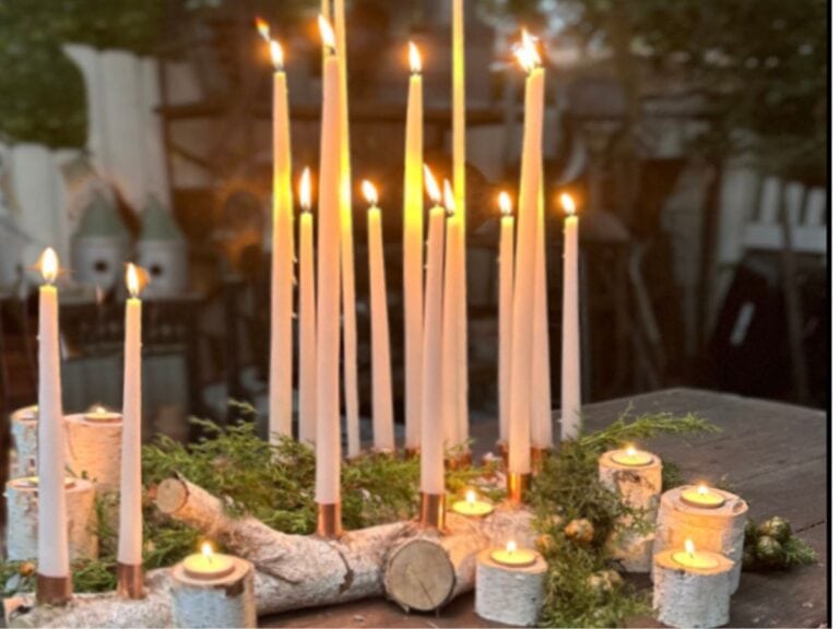 A collection of lit candles in various heights arranged on a wooden table. Set in DIY candle holders made from birch, greenery interspersed, they create a warm and cozy atmosphere. The softly blurred background hints at an inviting indoor setting.