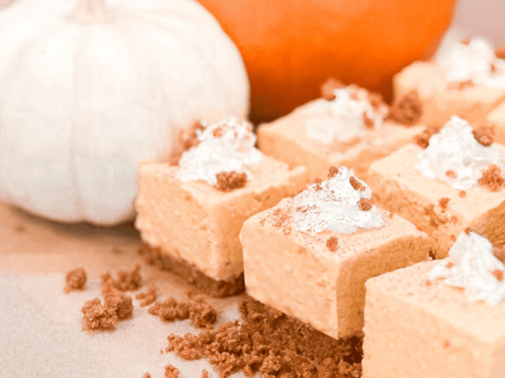 No Bake Pumpkin Cheesecake Bars topped with whipped cream and sprinkled with crumbs are arranged on a table. A white pumpkin and an orange pumpkin provide a charming autumn backdrop.