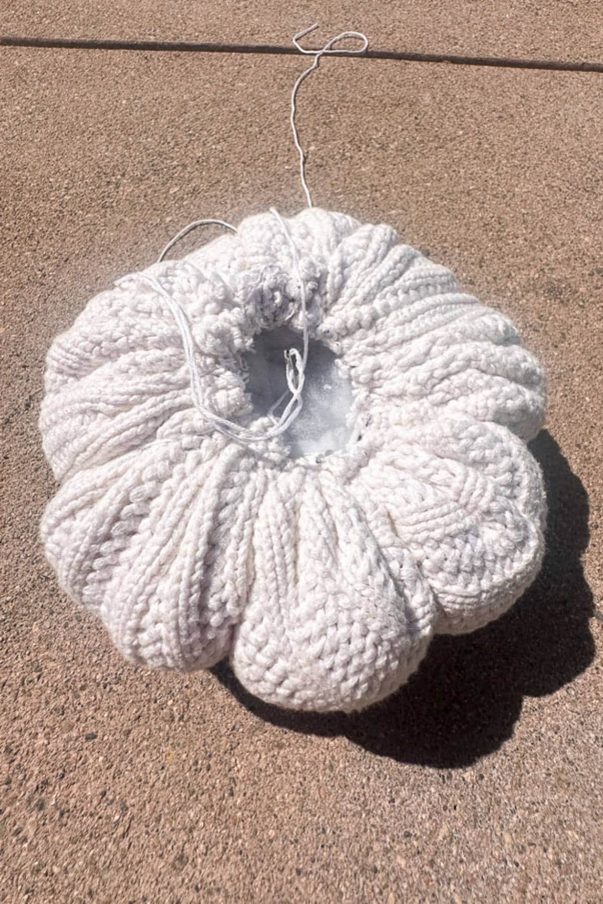 A knitted, off-white circular cushion is placed on a textured concrete surface. It has a gathered design in the center, with loose threads visible, suggesting it's a work in progress.