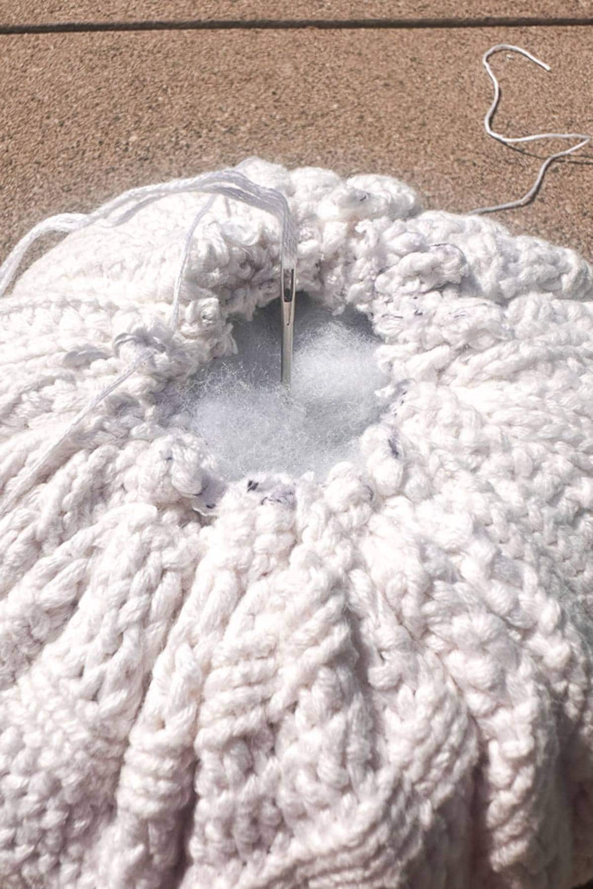 Close-up of a white knitted fabric being stitched closed with a needle and thread. The circular pattern is partially stuffed with white filling material, suggesting a work in progress, like a pillow or cushion, against a neutral background.