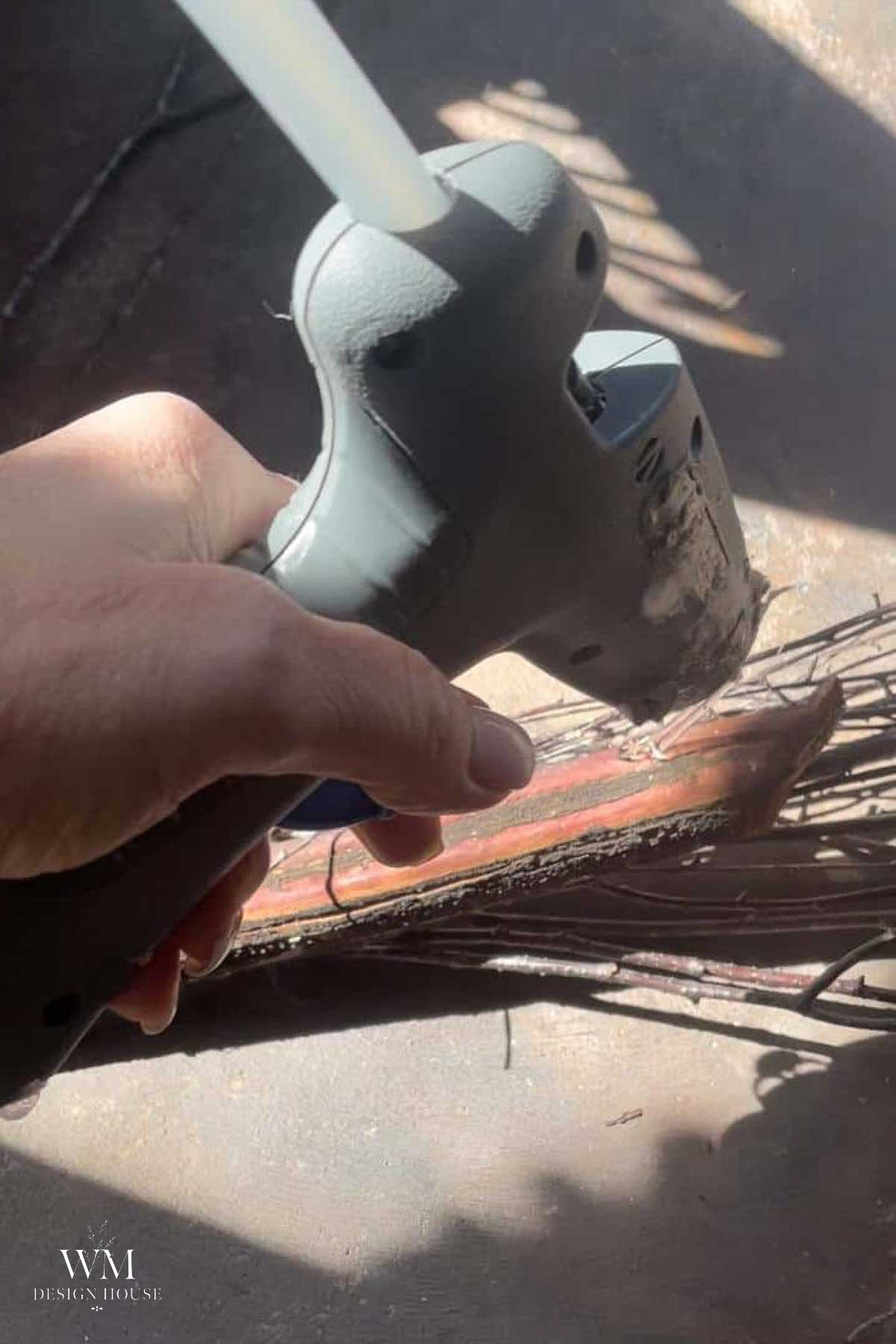 A hand uses a hot glue gun to apply glue to twigs and a red wooden stick, likely for a DIY Halloween decor project. Sunlight shines on the materials.