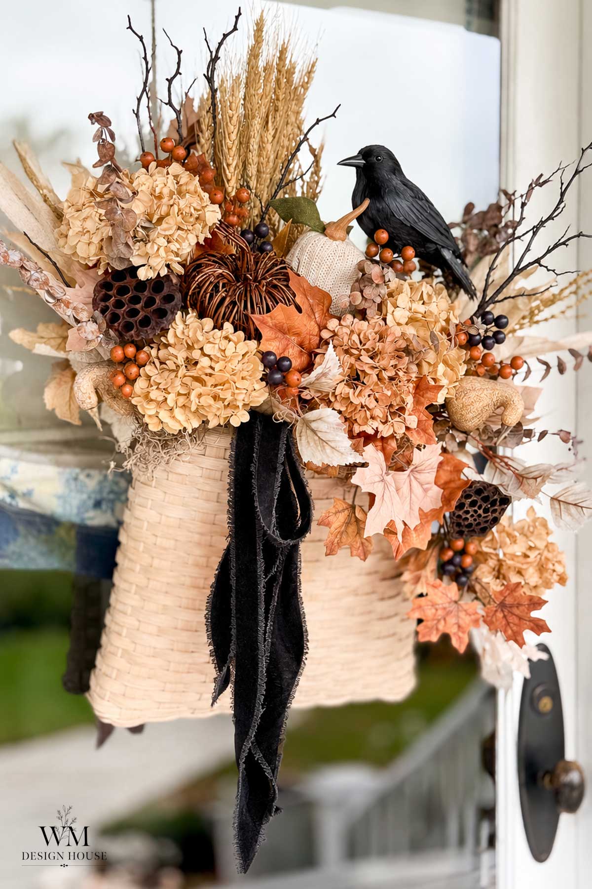 A woven basket with autumn flowers, berries, wheat, leaves, and a black crow hangs on a glass door. A black ribbon drapes from the arrangement, making this the perfect touch of DIY Halloween decor for your entryway.
