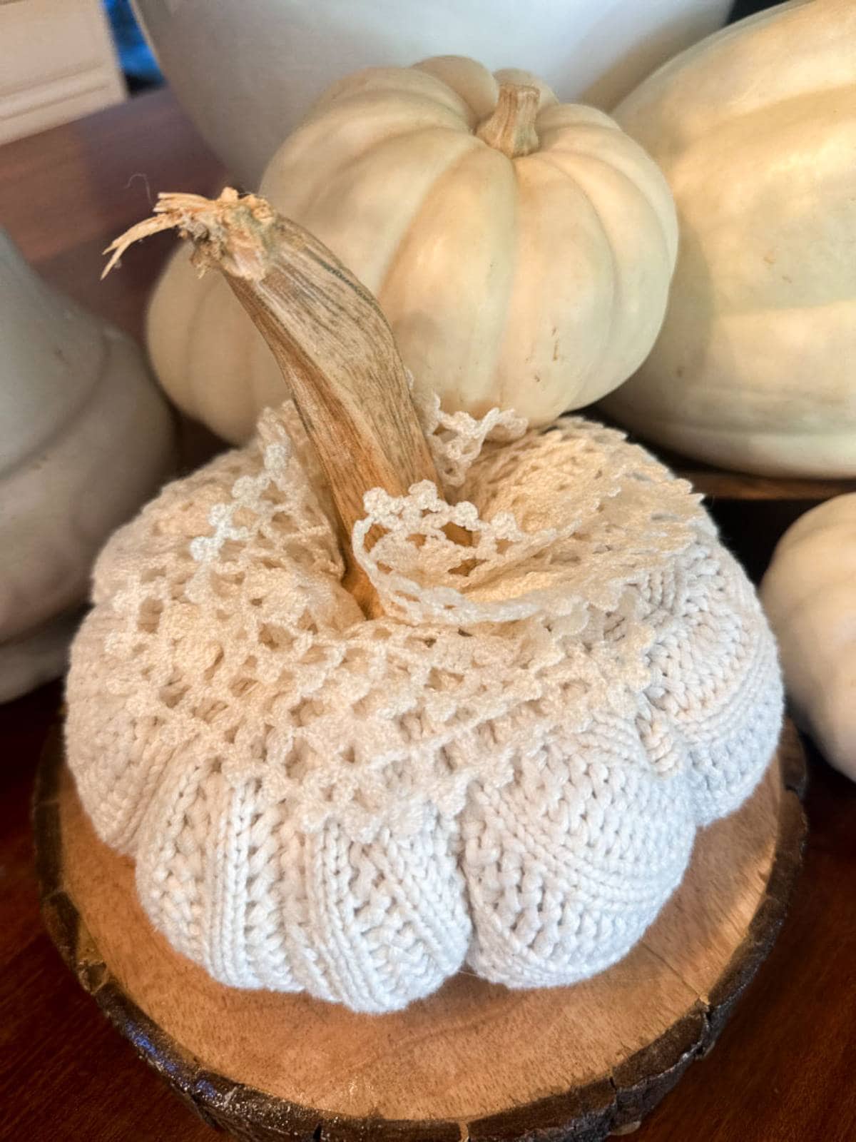 A small, cream-colored pumpkin decorated with a crocheted cover and lace sits on a wooden base. It's surrounded by other white pumpkins, creating a cozy, rustic autumn display.