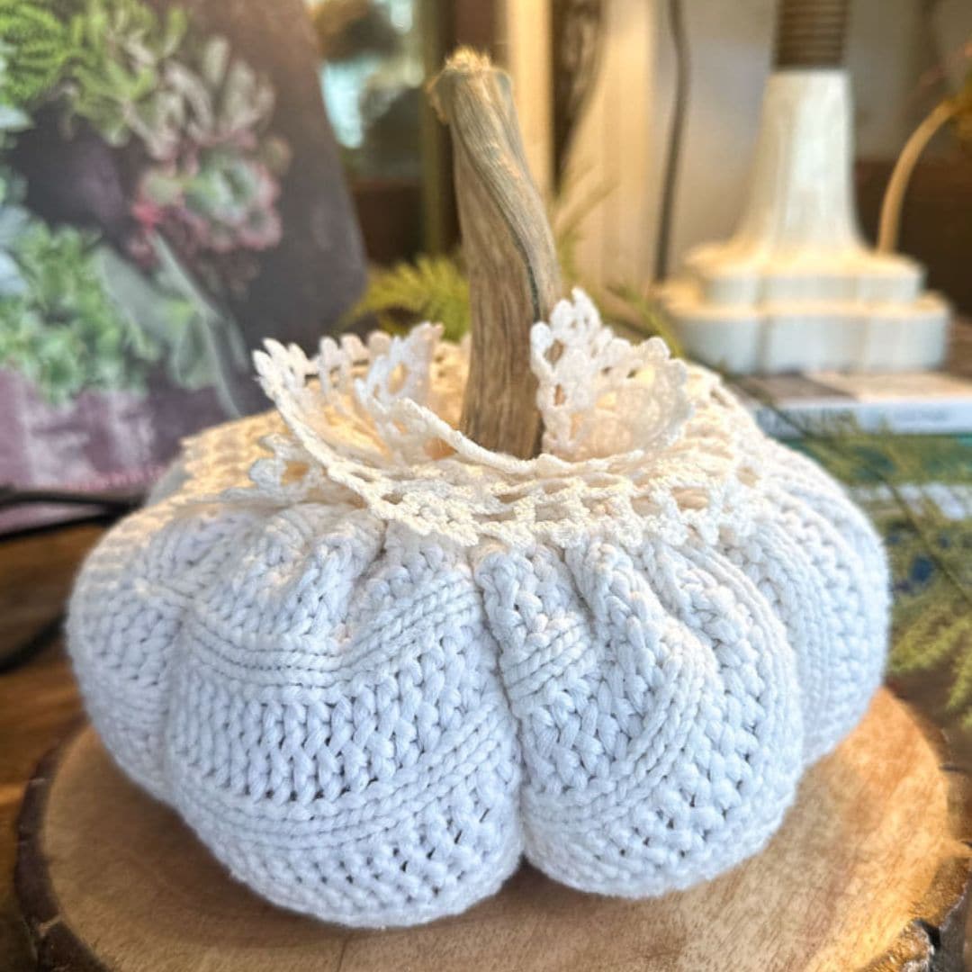 A knitted white pumpkin with a lace collar sits on a wooden surface, embodying the essence of September 2024. It boasts a natural-looking stem and is nestled in a cozy indoor setting, offering a perfect touch for seasonal charm.