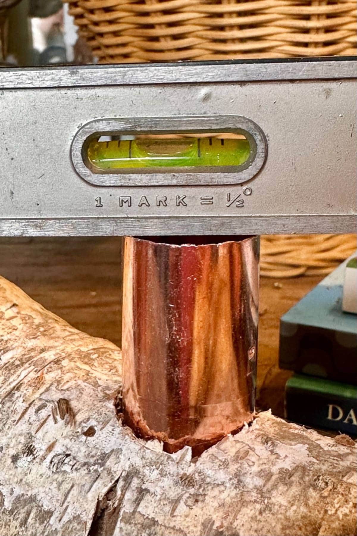 A close-up of a bubble level resting on a copper pipe, which is inserted into a branch. The bubble is centered, indicating a level position. In the background, there is a woven basket and part of a book visible.