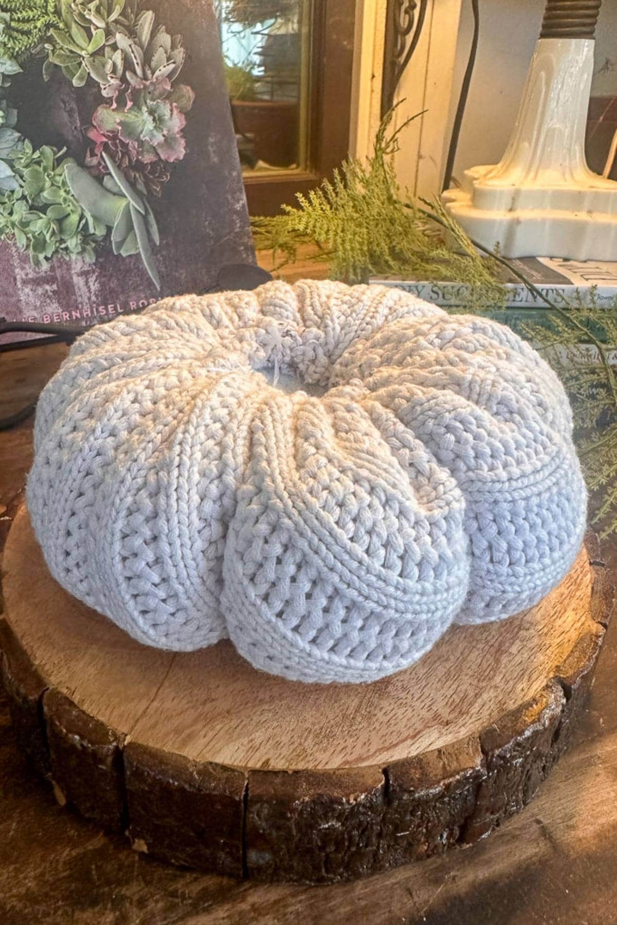 A light gray crocheted pumpkin sits on a circular wooden board. In the background, there is a lampshade and a partially visible book with floral imagery on the cover. Greenery decorates the scene.