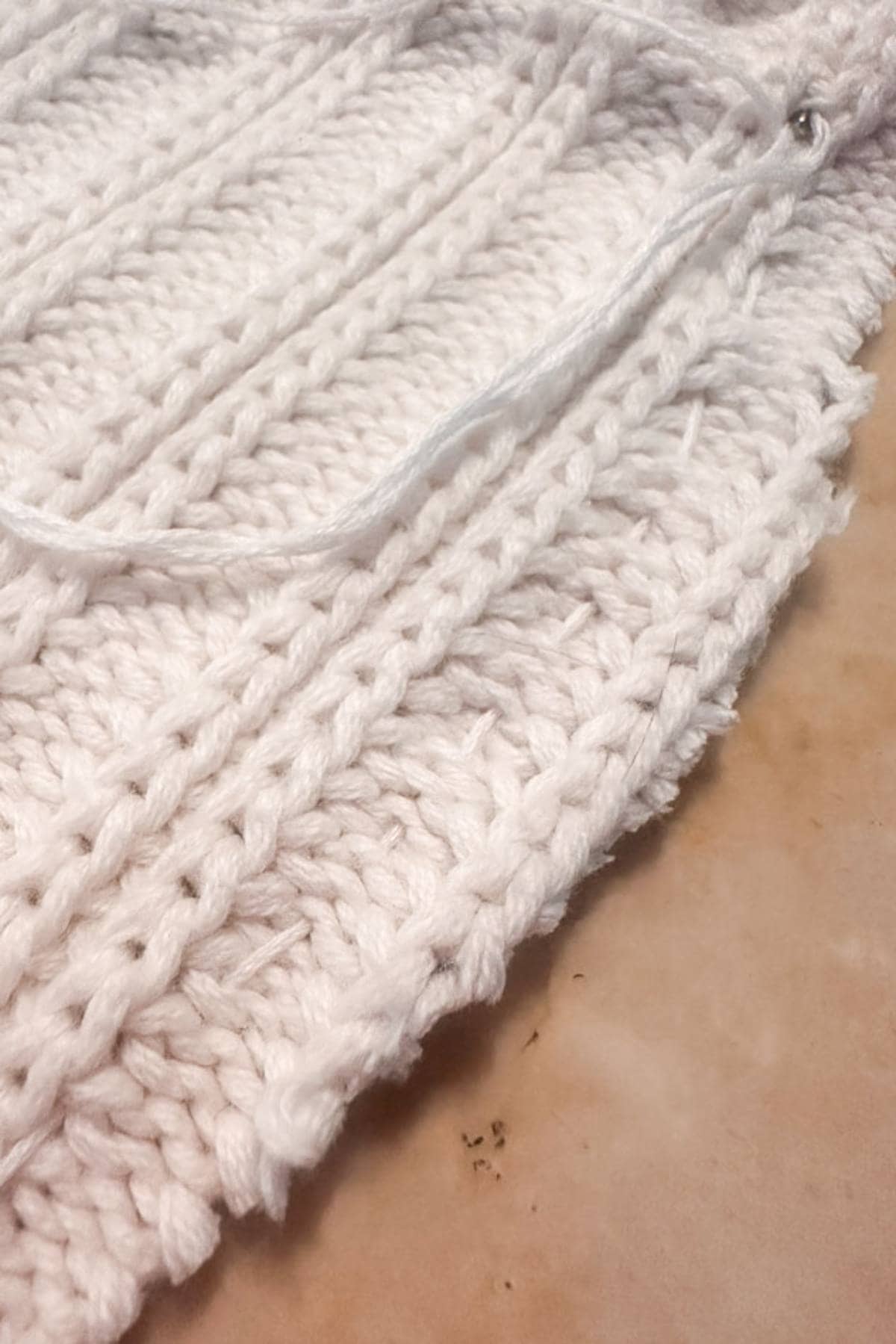 Close-up of white knitting sweater material with a running stitch. The knit pattern features vertical ribbed rows, with yarn strands visible on top.