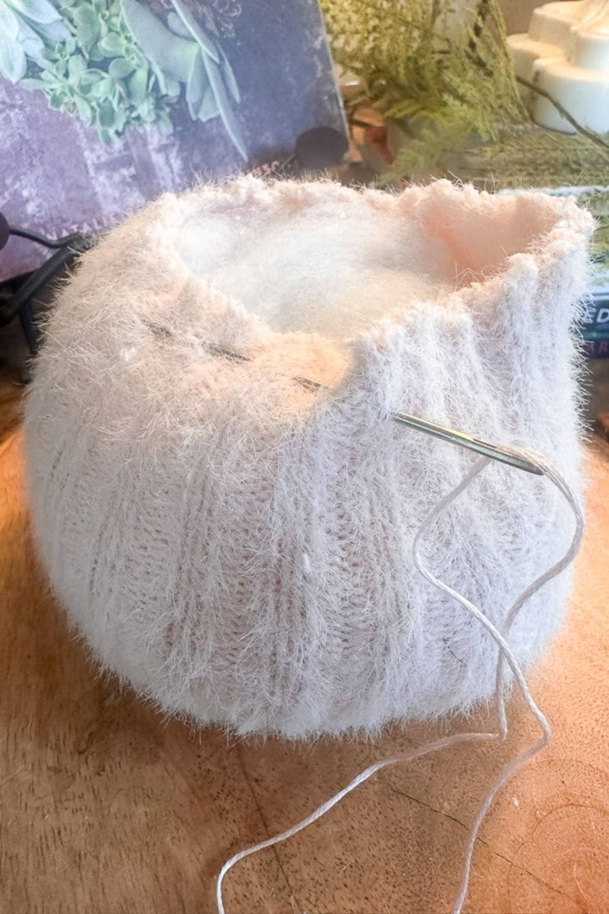 A fluffy, pastel pink knitted sweater pumpkin is being hand-stitched with a needle and white thread. It's resting on a wooden table with some crafting tools and materials in the background.