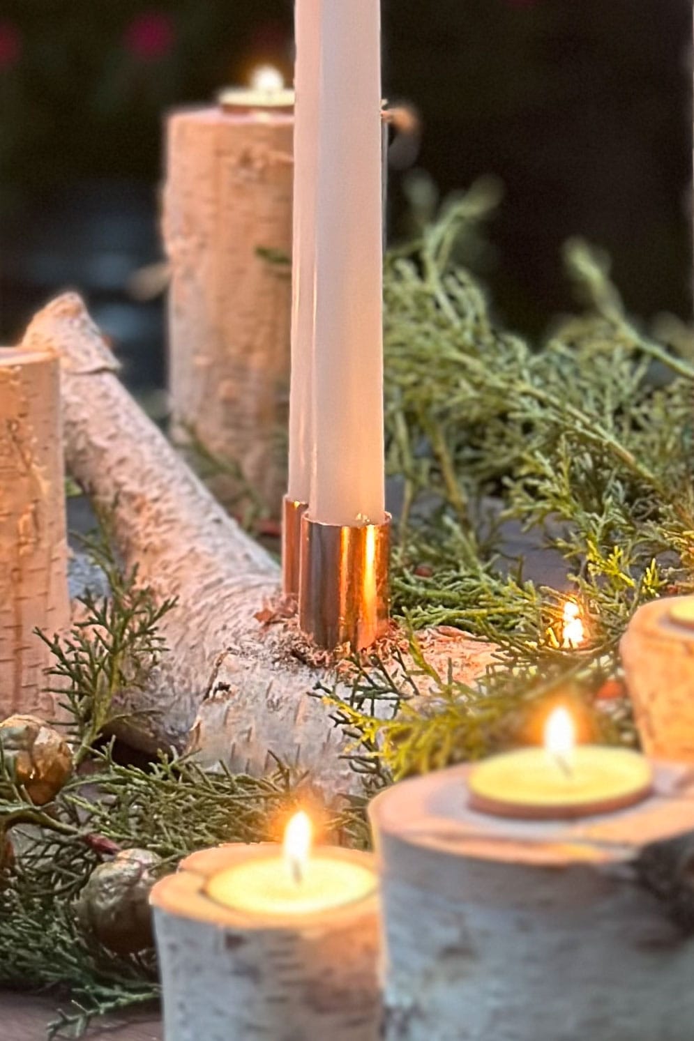 A tall white candle and several small tea lights are placed on rustic birch logs, doubling as charming DIY candle holders, surrounded by greenery. The scene is warmly lit, giving a cozy and festive atmosphere.