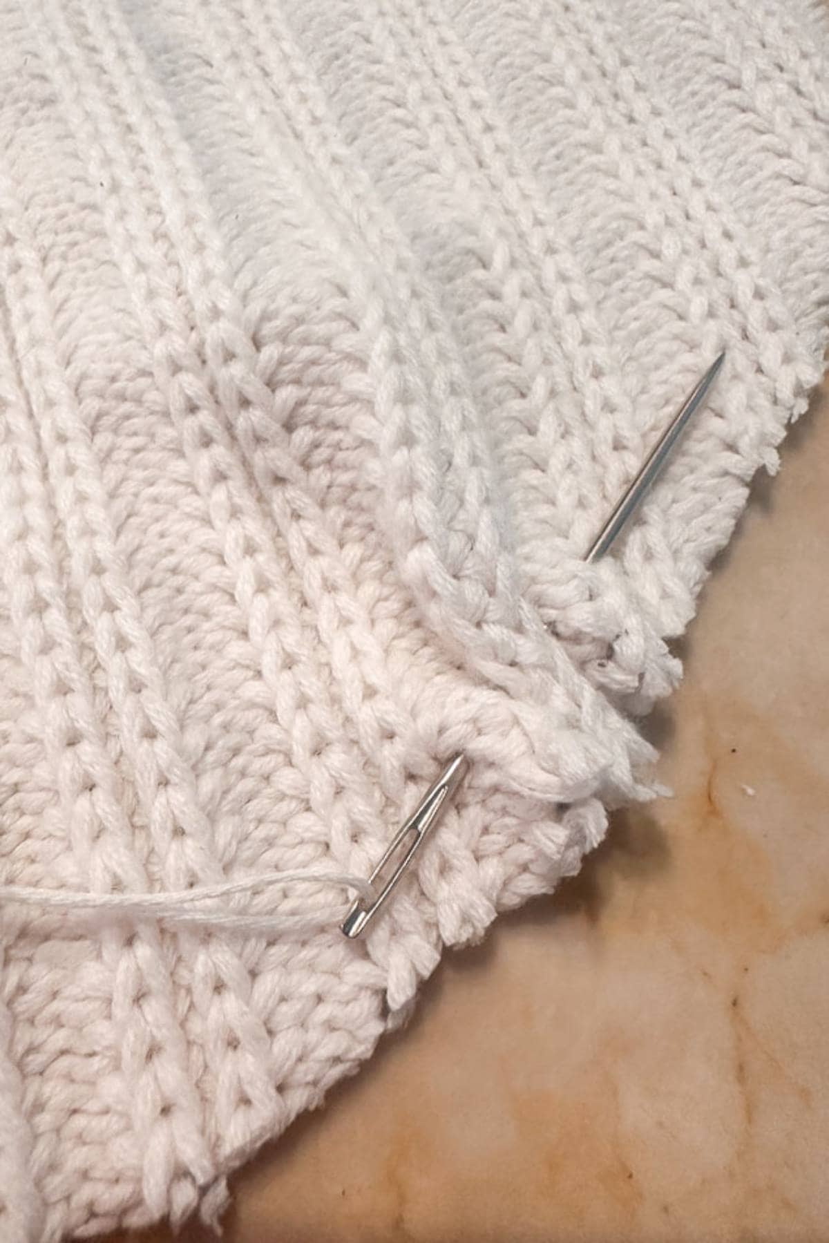 Close-up of white knit fabric with a ribbed pattern. Two metal needles are inserted into the knitting, and a loose yarn thread is visible. The background surface is a light-colored marble.