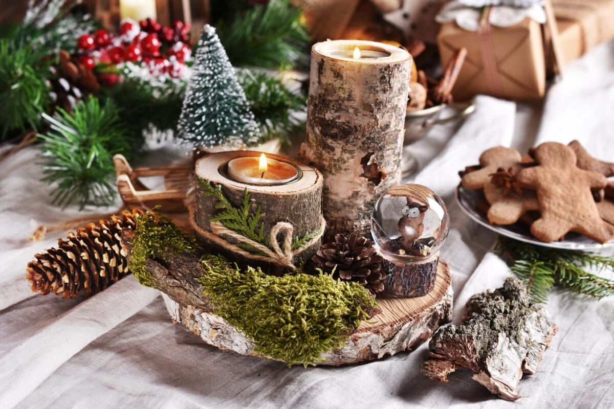 Create a rustic holiday centerpiece with DIY candle holders crafted from birchwood featuring two lit candles. This charming display includes a small snow globe, pine cones, and moss on a wooden base, adorned with a miniature tree and surrounded by festive greenery and gingerbread cookies.