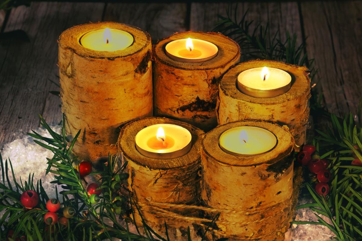 Five lit candles are nestled inside rustic birch log holders, surrounded by evergreen branches and red berries, set against a wooden background, creating a warm and festive atmosphere.