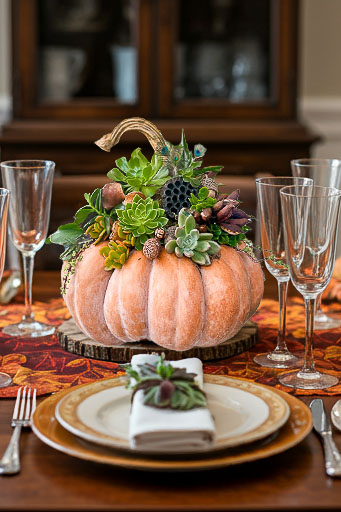 A decorative centerpiece made of a light orange pumpkin with various succulents arranged on top. It sits on a wooden slice, surrounded by elegant tableware, including plates, utensils, and crystal glasses on a fall-themed tablecloth.