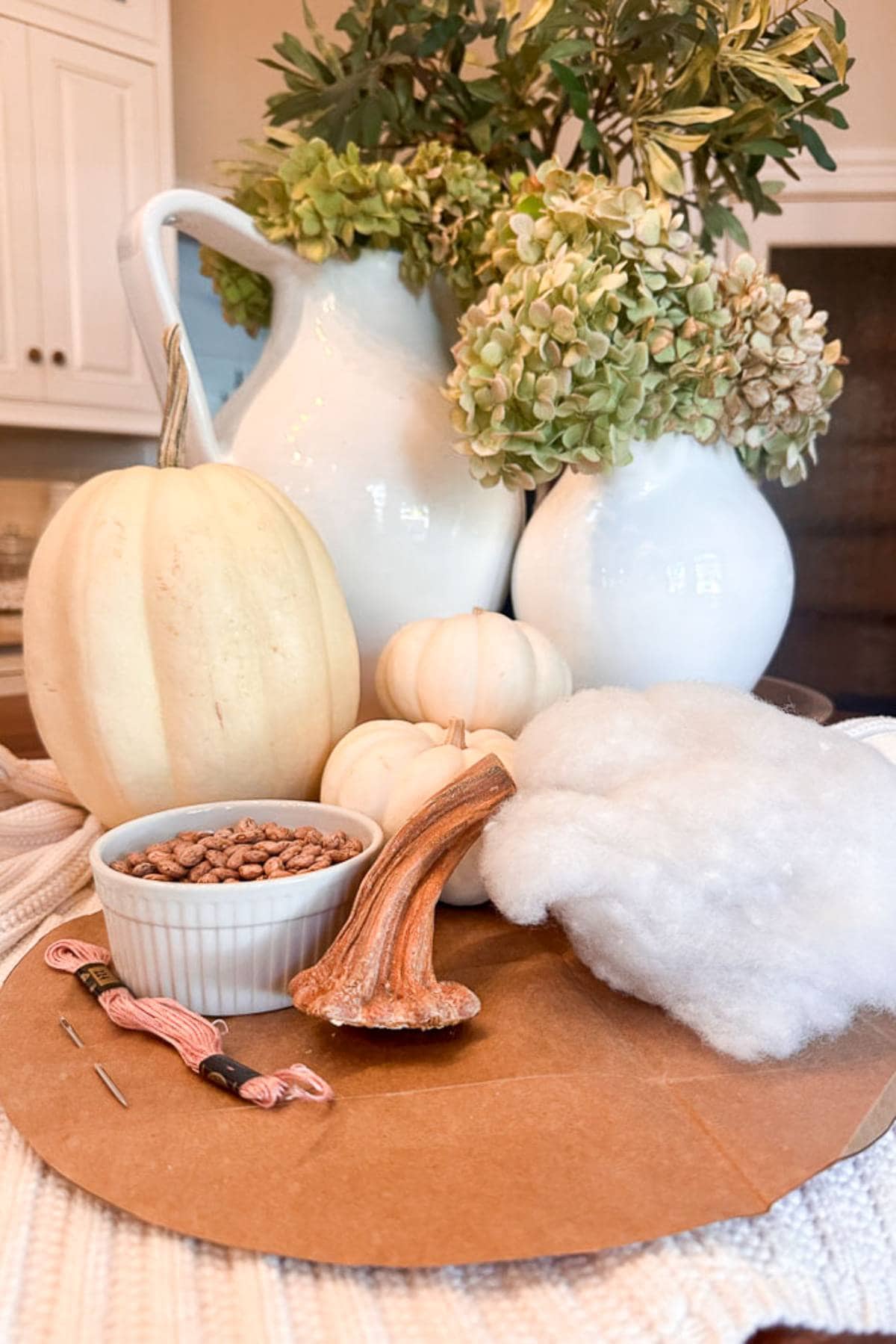 A crafting scene with materials for a fall project: two white ceramic pitchers with hydrangeas, a large white pumpkin, two small pumpkins, a bowl of seeds, embroidery thread, fabric, and a pumpkin stem on a round cardboard base.