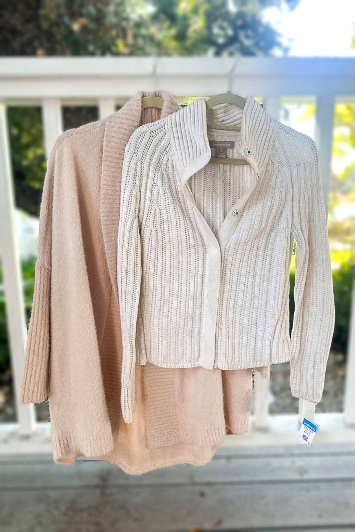 Two beige knit sweaters hang on hangers outdoors. One is a button-up with a collar, and the other is an open-front cardigan. The background features a white railing and blurred greenery.