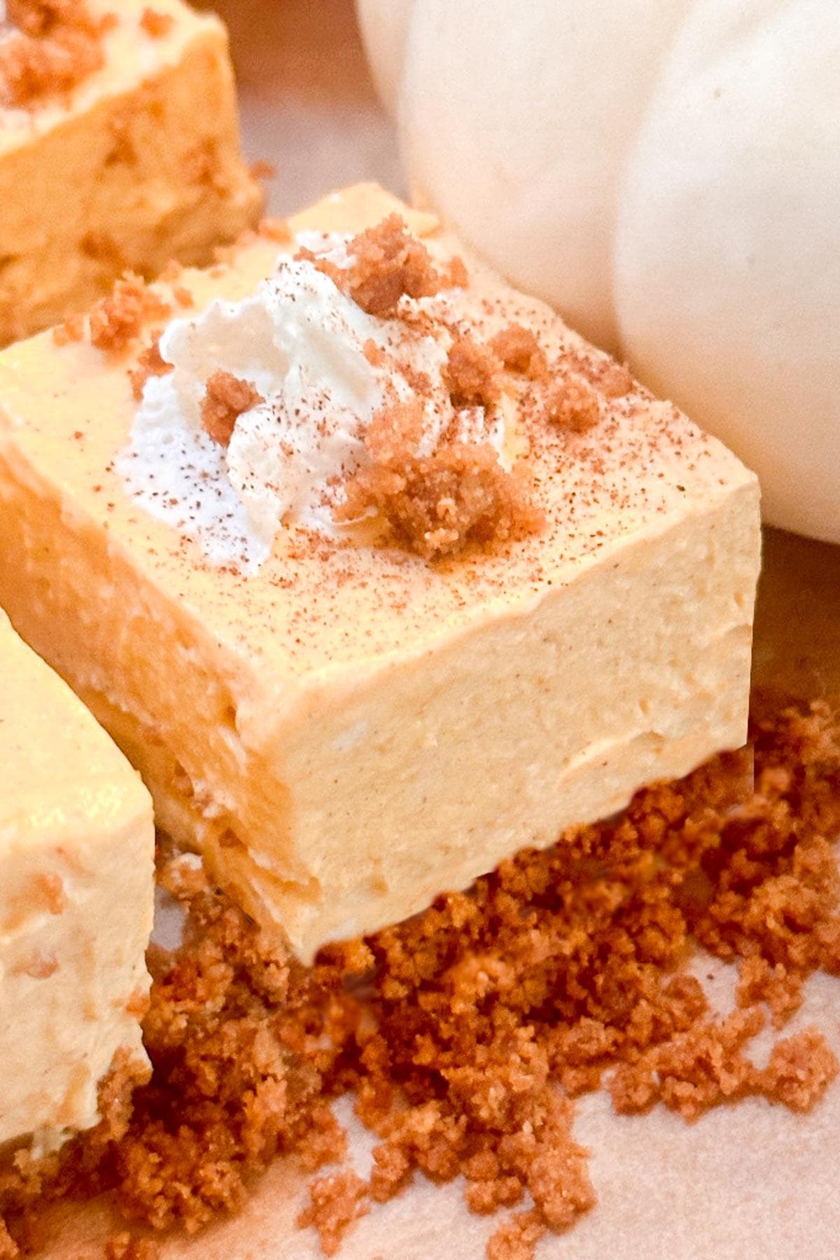 A square piece of no bake pumpkin cheesecake bars topped with whipped cream and crumbled graham crackers. It's surrounded by more crumbs, with a blurred white pumpkin in the background.