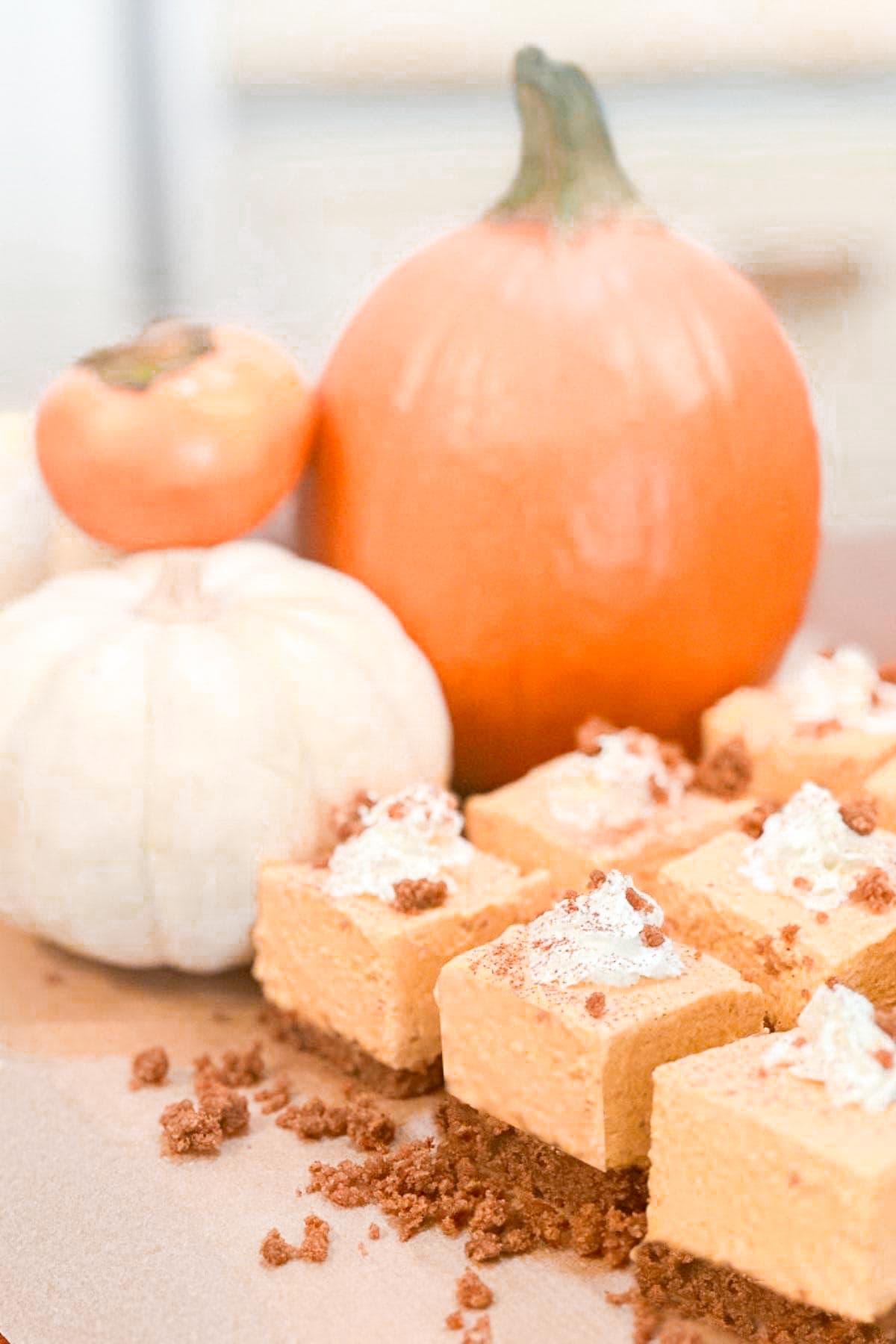 No Bake Pumpkin Cheesecake Bars, topped with whipped cream and sprinkled with crumbs, are displayed beside two pumpkins—one orange and one white—on a wooden surface.