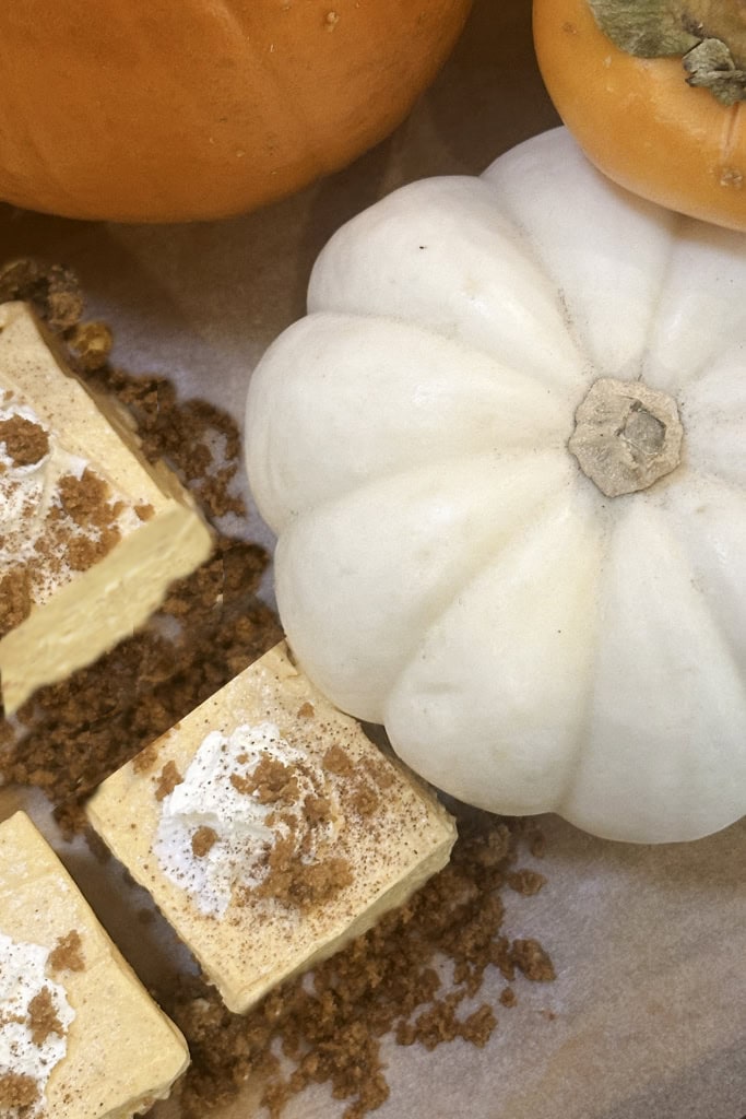 Three no-bake pumpkin cheesecake bars with crumbly topping sit beside a white pumpkin and a small orange gourd on parchment paper. The autumn-themed dessert arrangement highlights the flavors of the season.