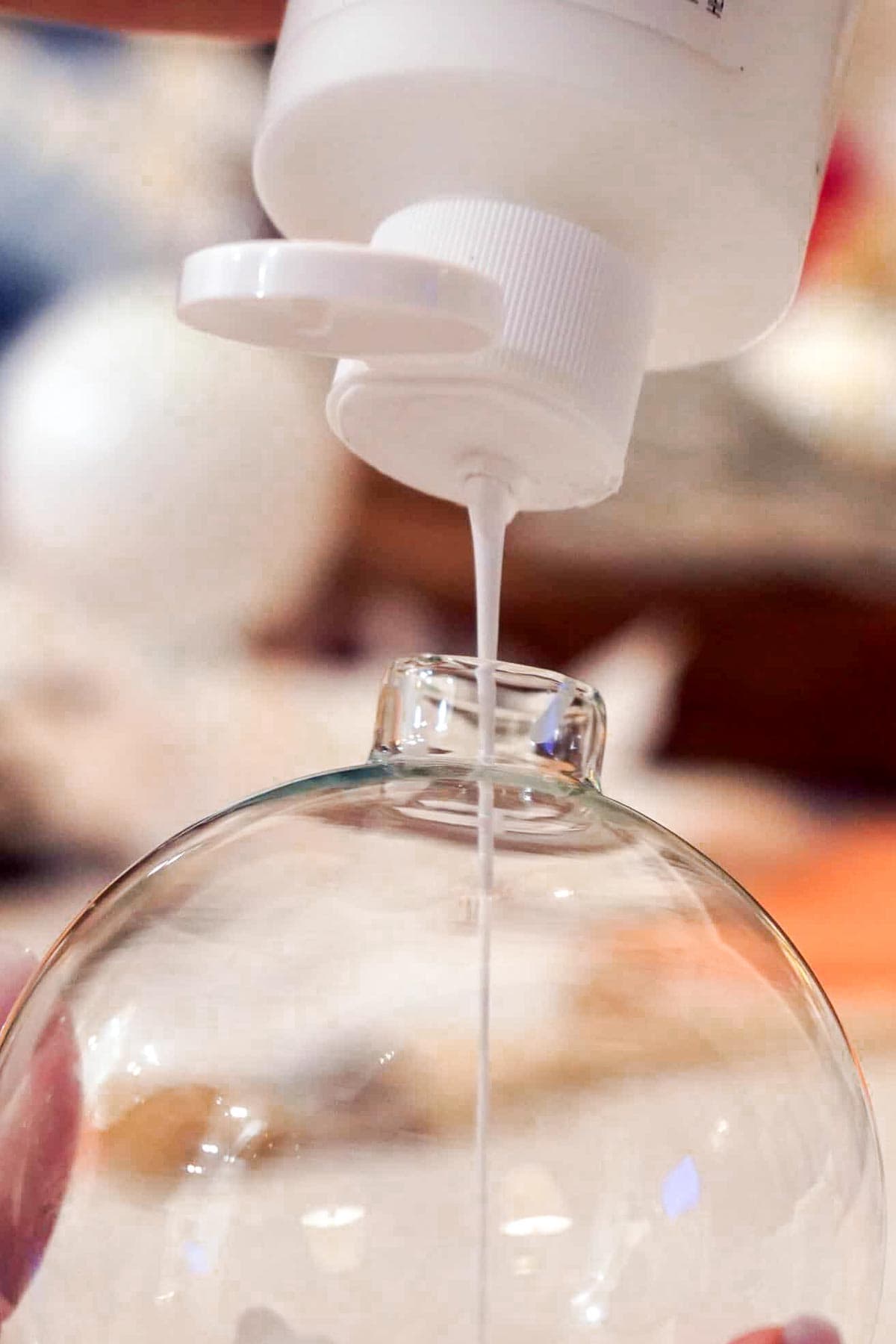 A person fills a clear glass ornament with white liquid glue from a squeeze bottle. The ornament is held gently, and the glue flows into the open neck of the ornament. The background is blurred, focusing on the crafting activity.