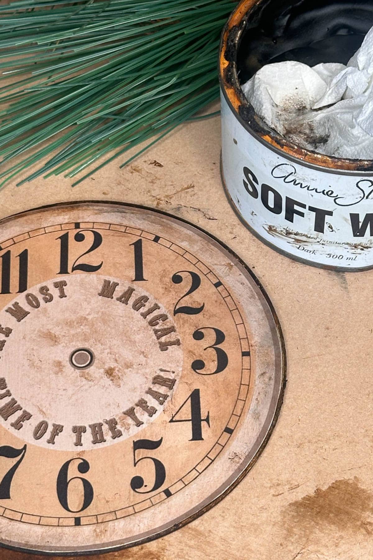 A wooden clock face, adorned with numbers and the phrase "The Most Magickal Time of the Year," rests on a table beside an open can labeled "Soft Wax," stuffed with paper towels. Pine needles peek from the background, evoking a scene perfect for crafting DIY vintage Christmas ornaments.