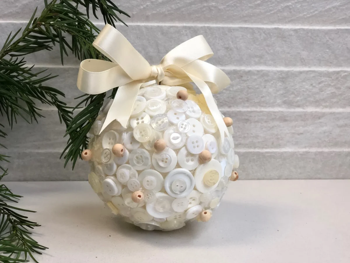 A DIY vintage Christmas ornament, elegantly covered in white buttons and small wooden beads, is adorned with a cream ribbon bow on top. It rests on a white surface beside pine branches, against a light-textured background.