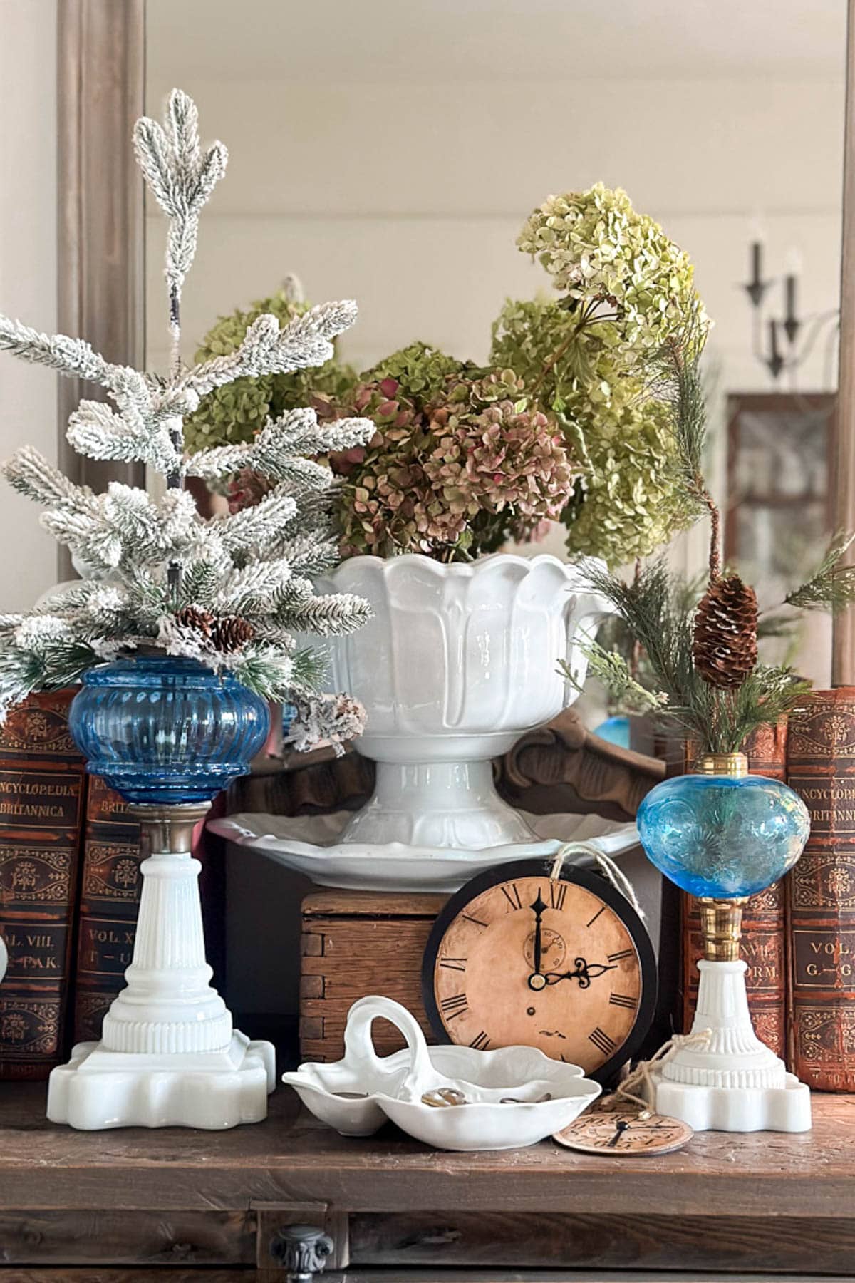 A decorative arrangement on a shelf features vintage books, DIY vintage Christmas ornaments, a small white ceramic Christmas tree, hydrangeas in a vase, blue glass lamps, a wooden clock, and a dish with buttons. A mirror reflects the items, adding depth to the display.