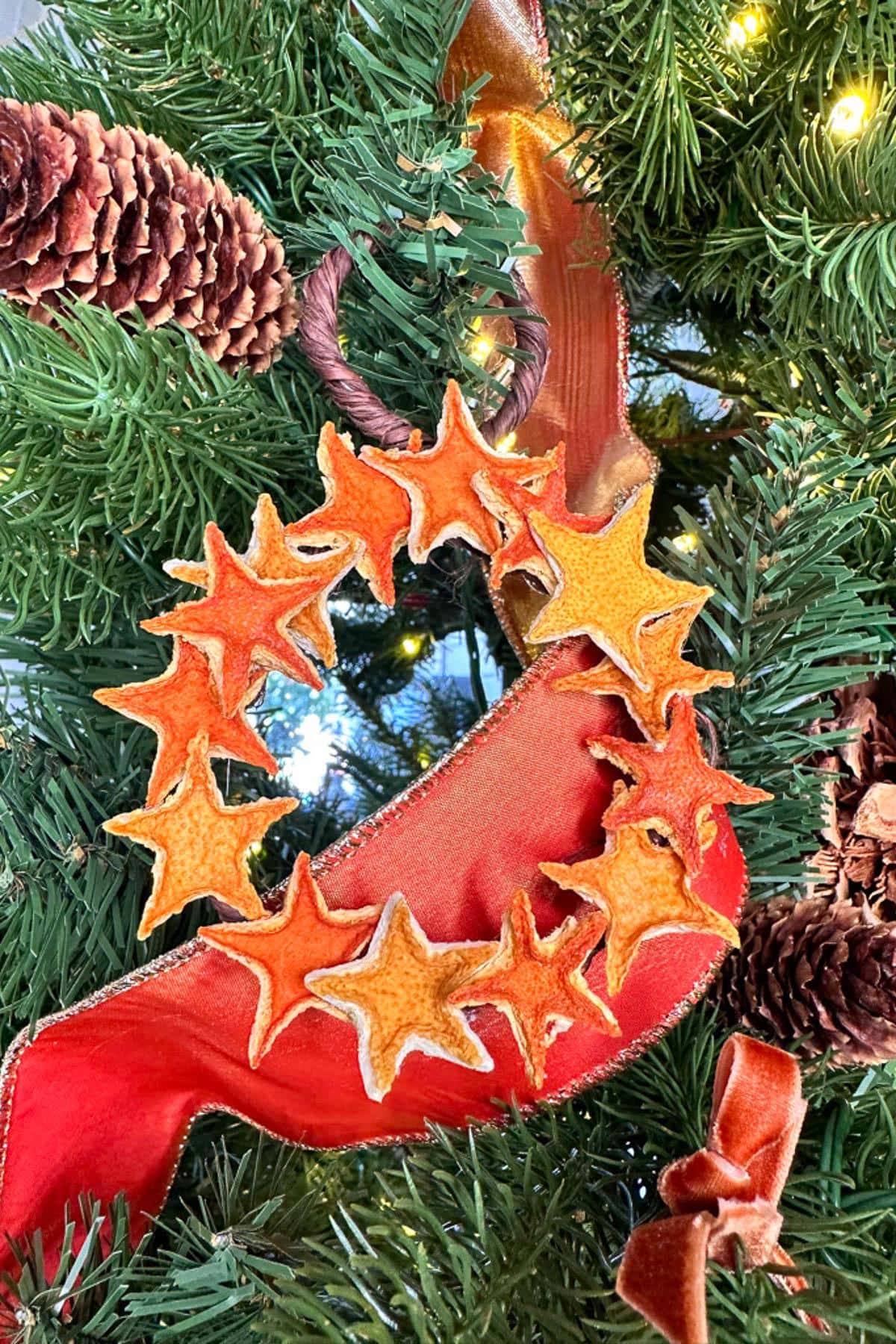 A festive wreath made of orange and yellow star-shaped decorations and a red ribbon adorned with pinecones hangs on a green Christmas tree.