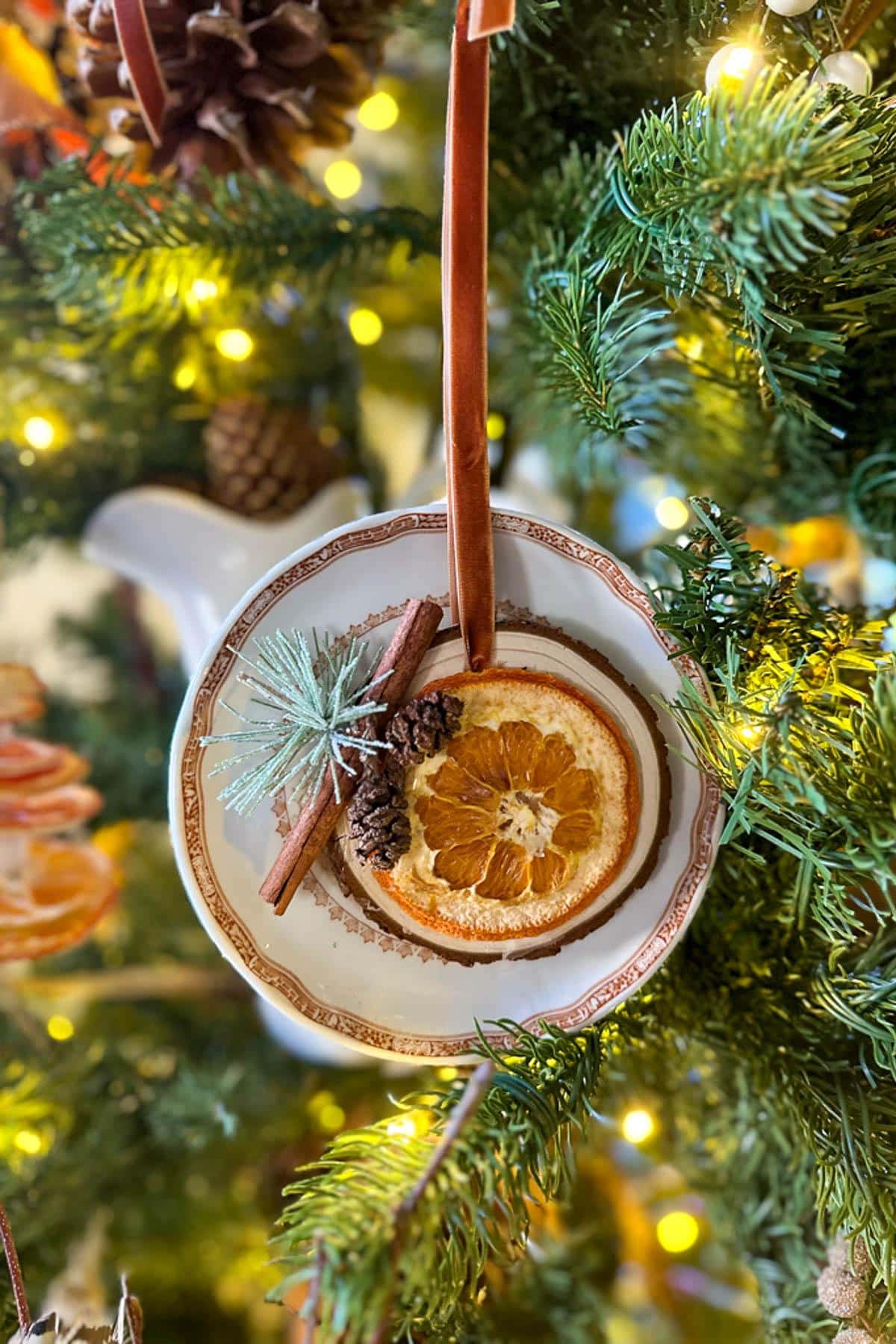 Festive decoration hanging on a Christmas tree branch, featuring a teacup filled with dried orange slices, cinnamon sticks, and a pinecone, surrounded by twinkling lights and green foliage.