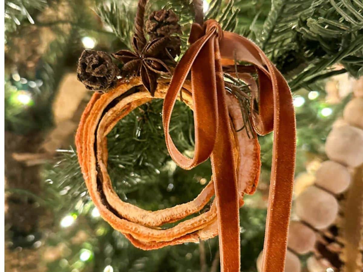 Close-up of a handcrafted dried orange ornament featuring cinnamon sticks, tied with a brown ribbon. It hangs gracefully on a Christmas tree adorned with twinkling lights.