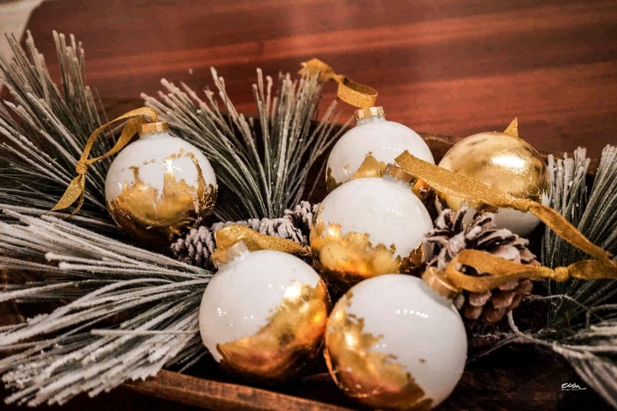 A wooden tray holds white and gold Christmas ornaments adorned with gold ribbons. The ornaments rest on frosted pine branches and pinecones, creating a festive decoration on a wooden surface.