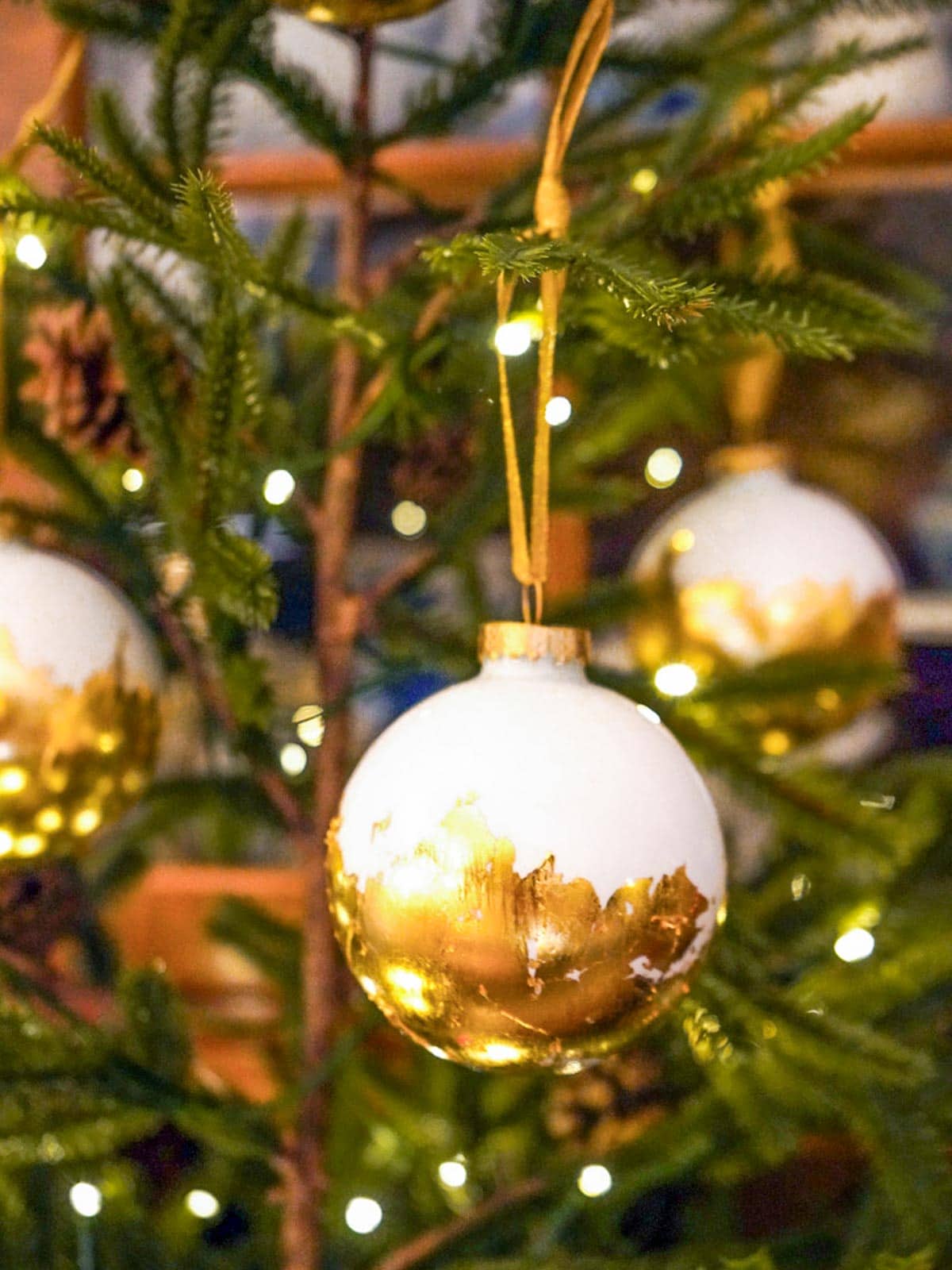 A close-up of a Christmas tree with white and gold baubles hanging from the branches. The tree is lit with small, warm white lights, creating a festive and cozy atmosphere.