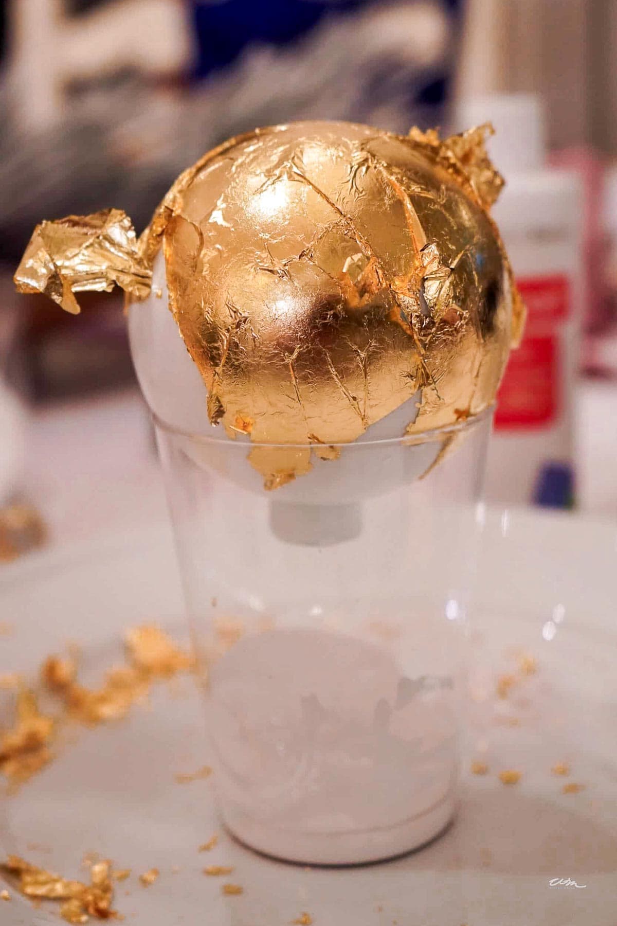 A light bulb partially covered in gold leaf rests on a plastic cup. Torn pieces of gold leaf are scattered around on the surface. A red and white bottle is visible in the blurred background.