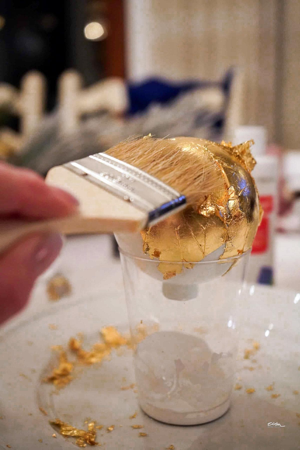 A hand uses a brush to apply gold leaf onto a spherical ornament placed on a clear plastic cup. Gold leaf fragments are scattered around the ornament, creating a decorative and artistic process.