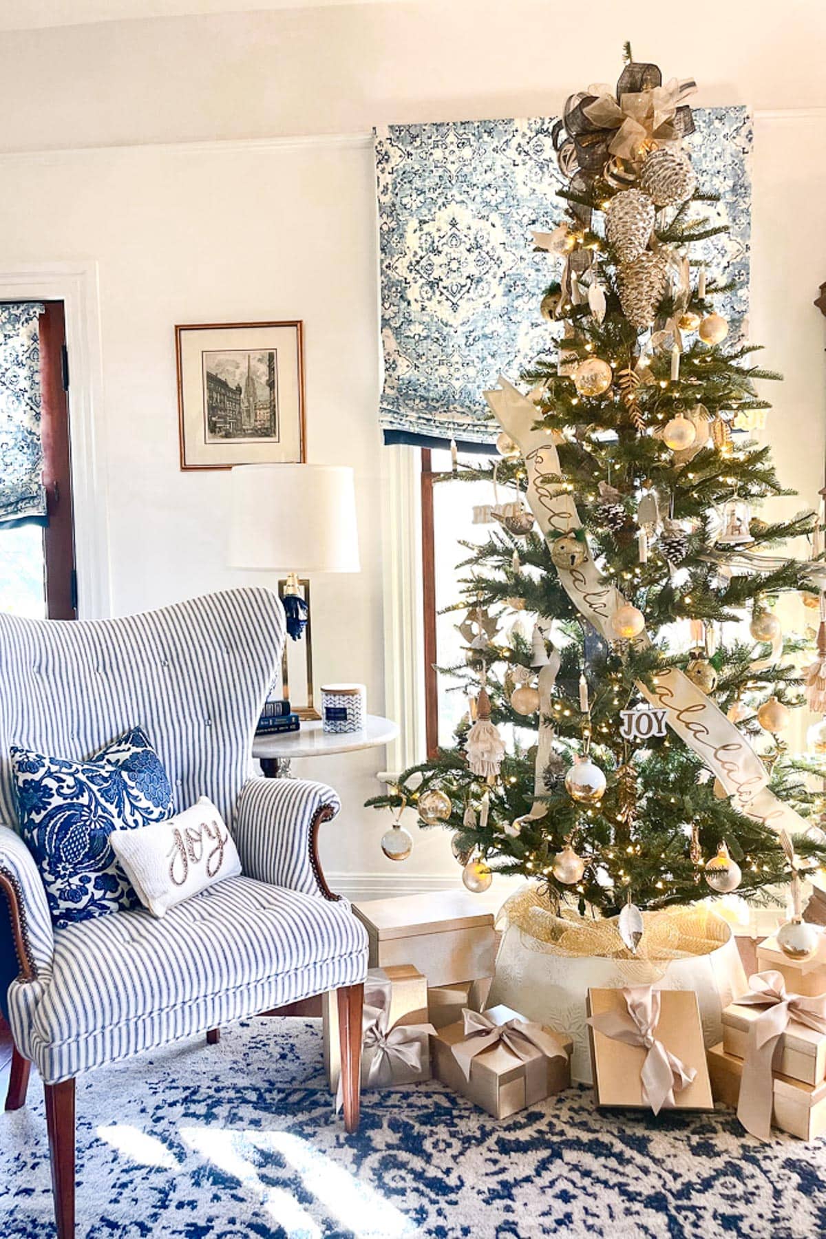 A cozy living room with a Christmas tree adorned with ornaments and ribbons next to wrapped gifts. A striped armchair with "Joy" pillow sits to the left, near a window with patterned curtains. A table lamp and framed pictures complete the decor.