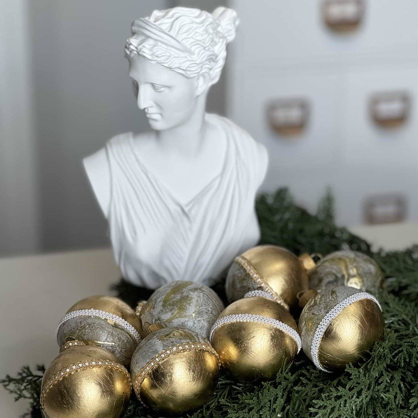 A white marble bust surrounded by decorative gold and silver Christmas ornaments, with greenery at the base. The background features a soft-focus white interior with square wall accents.