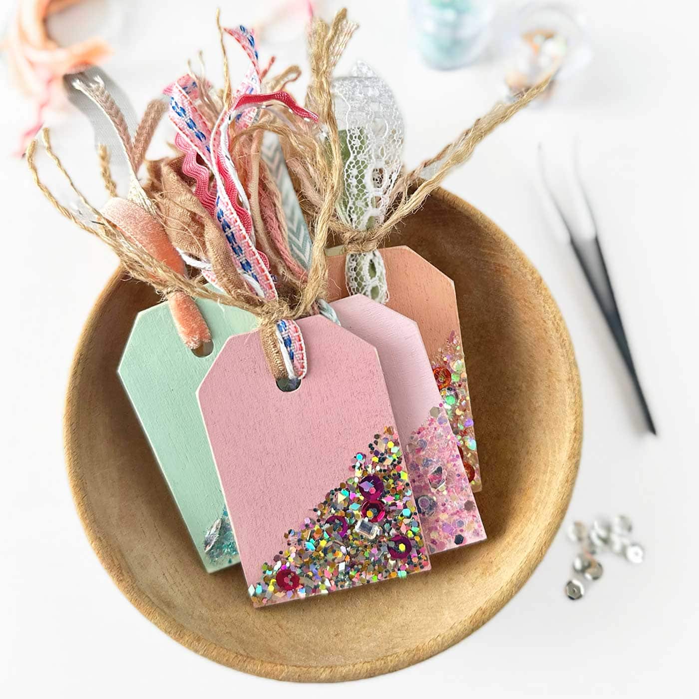 A wooden bowl contains four decorative gift tags, each adorned with colorful glitter and lace ribbons. The tags are painted in pastel shades of pink and mint green. Scissors and loose rhinestones are partially visible on the table.