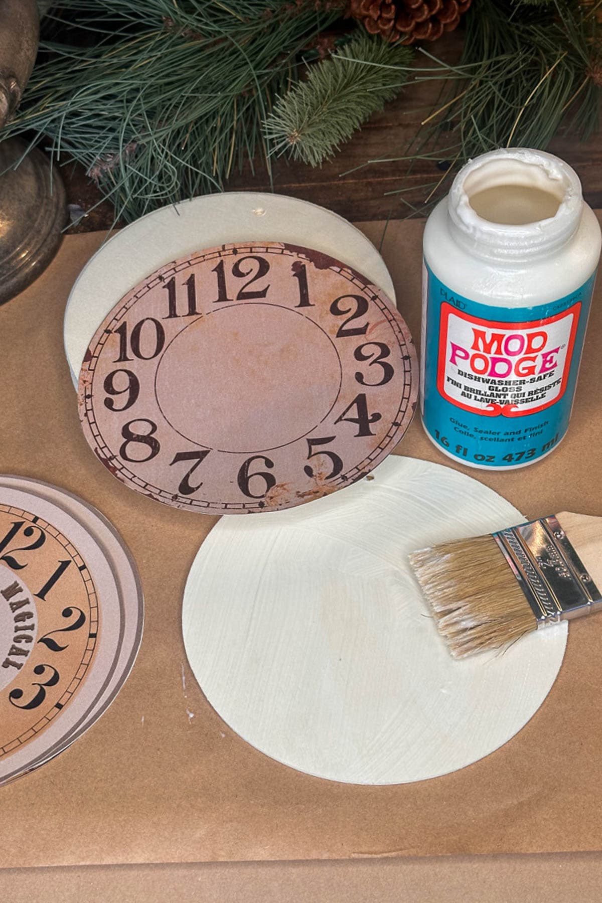 A crafting scene features a DIY vintage Christmas ornament with a painted circle like a clock face, paired with Mod Podge glue, a brush, and paper adorned with printed numbers. Pine branches create a festive backdrop for this nostalgic creation.