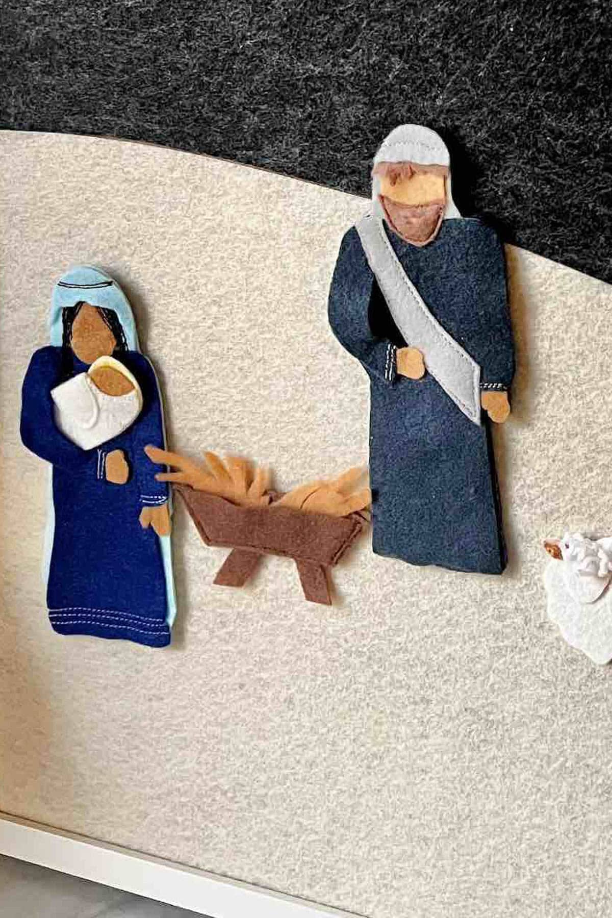 This felt artwork captures the warmth of a nativity scene, where figures of a woman with a baby and a man gather around a manger filled with hay, evoking the cozy spirit of a Christmas pajama party.