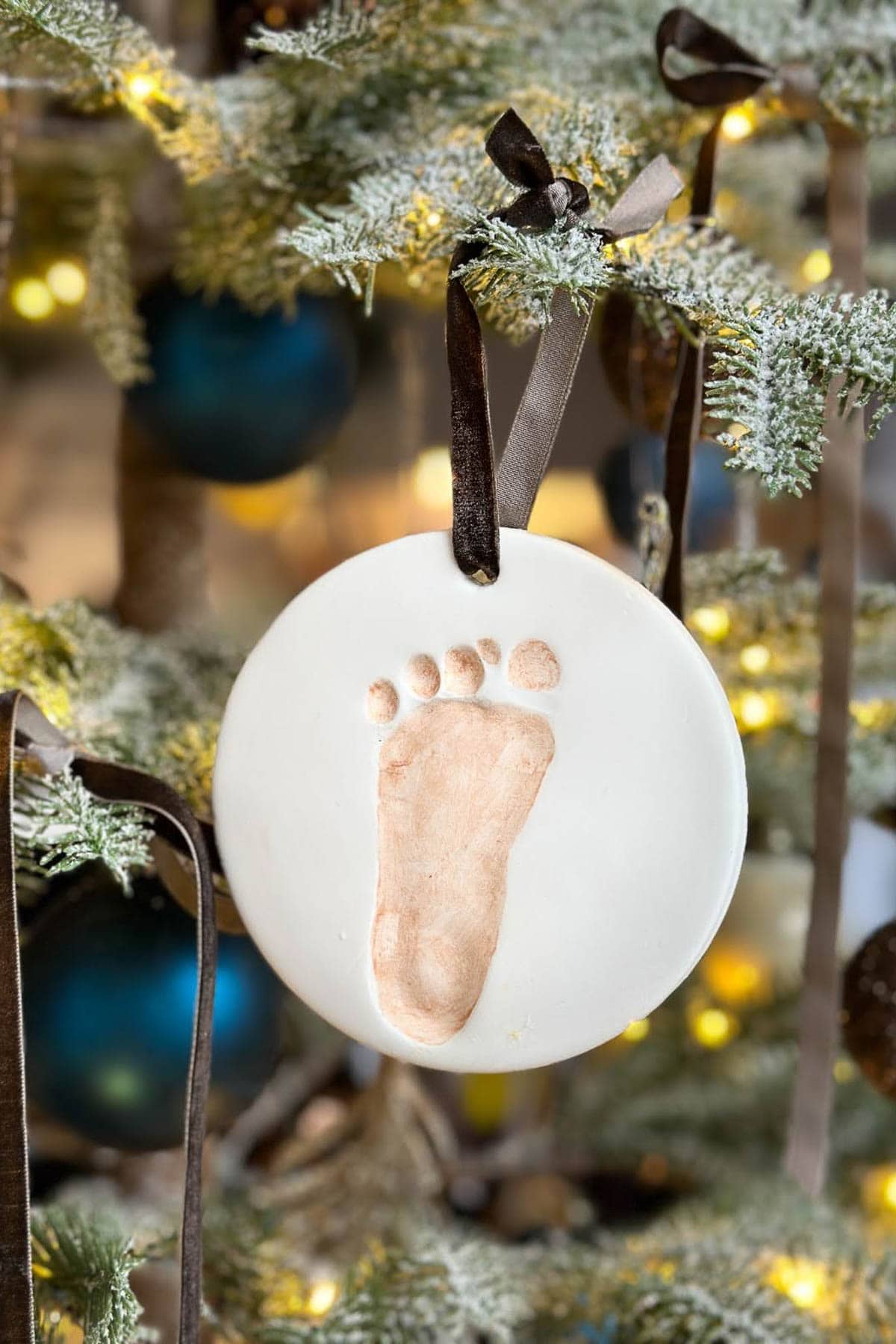 A round white ornament with a small footprint imprint, reminiscent of a joyful Christmas pajama party, hangs from the tree. The light brown footprint is complemented by a dark ribbon. The tree sparkles with blue baubles and glowing lights, setting a festive scene.