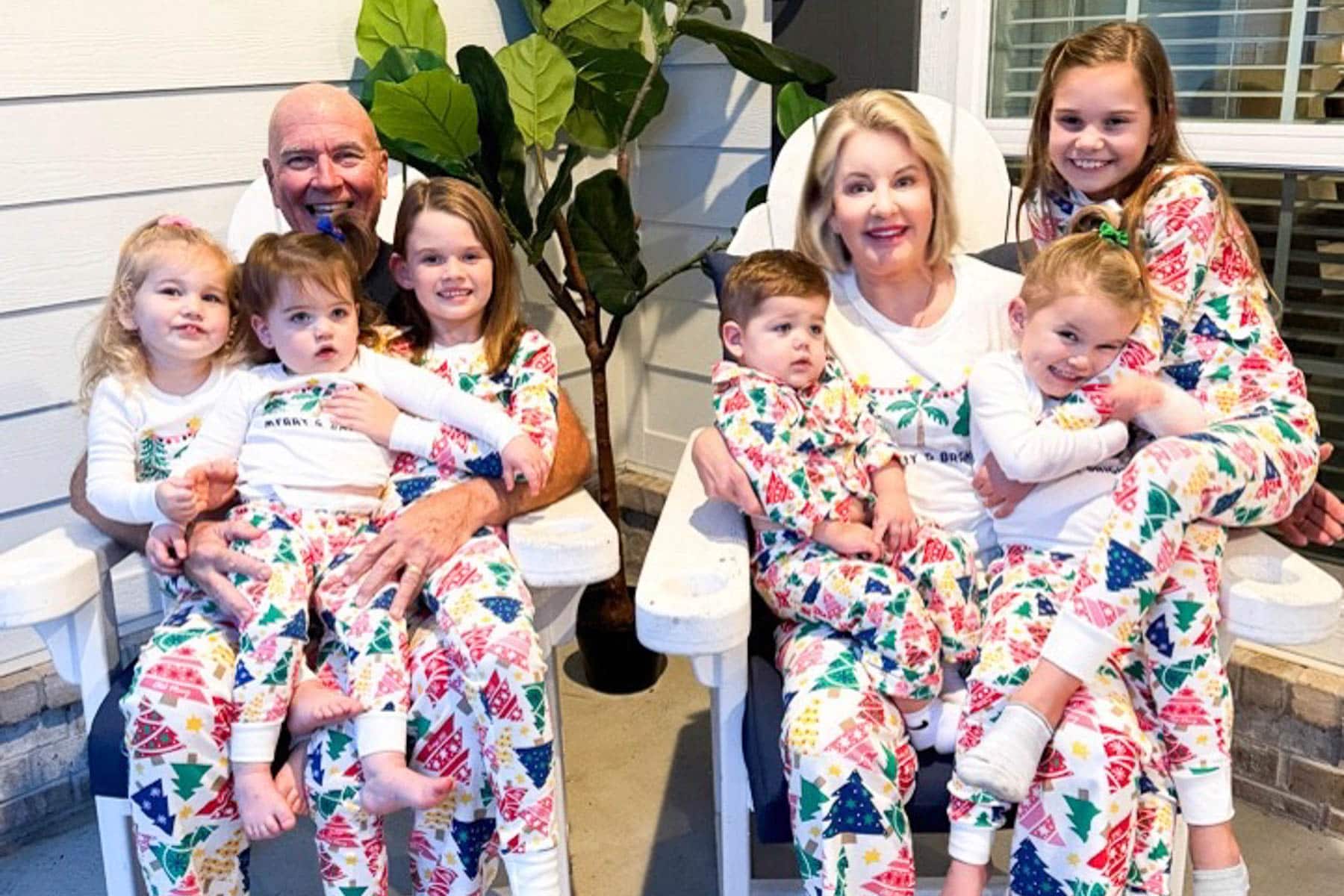 A family poses on a porch in matching Christmas-themed pajamas, capturing the joy of their festive pajama party. Two adults sit on chairs, surrounded by six smiling children. A potted plant adds a touch of greenery in the background, as everyone beams with holiday cheer.