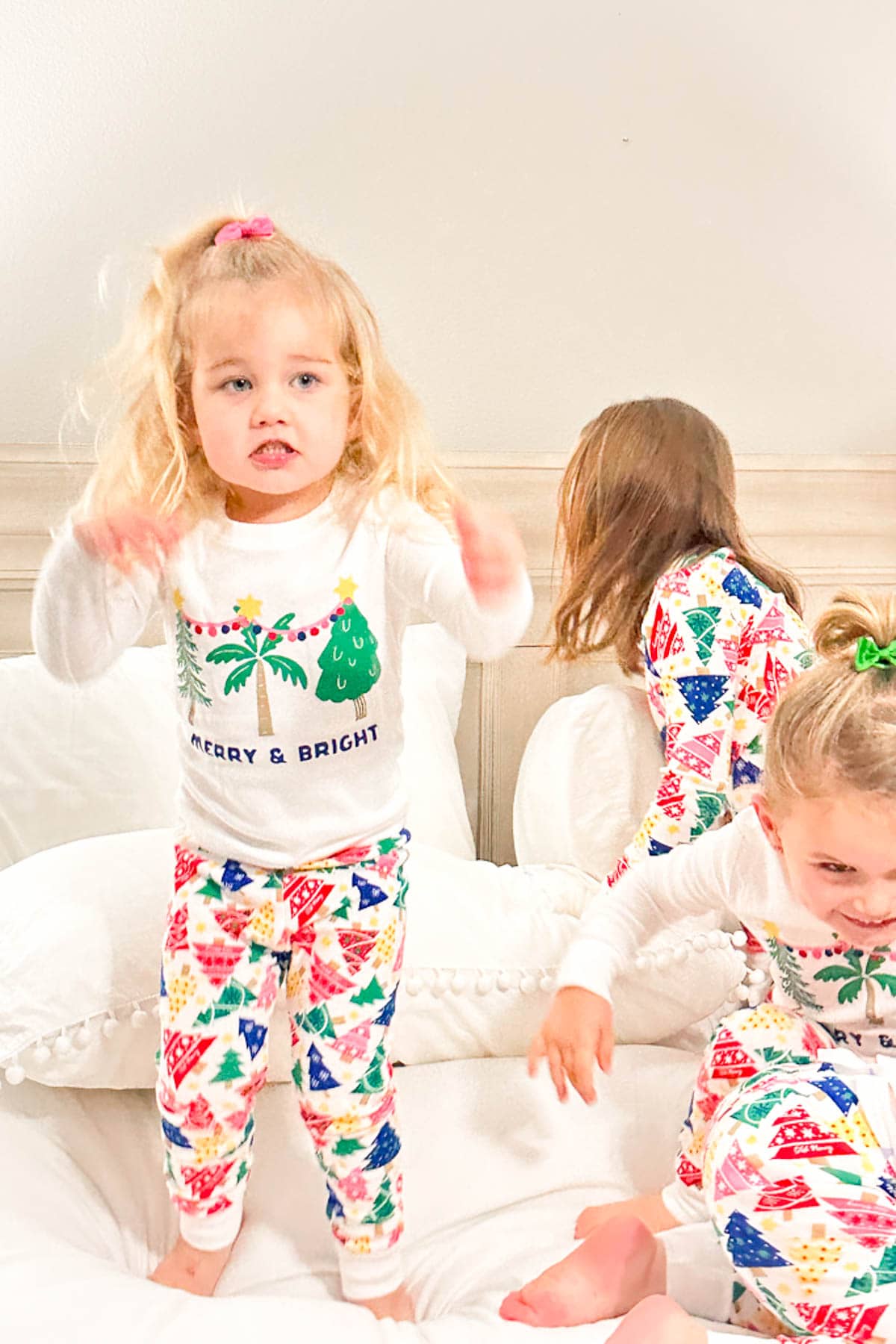 Three young grandkids in festive pajamas with colorful Christmas tree patterns are playing on a bed. One child faces the camera while the others are turned away, creating a lively and joyful Christmas pajama party atmosphere.