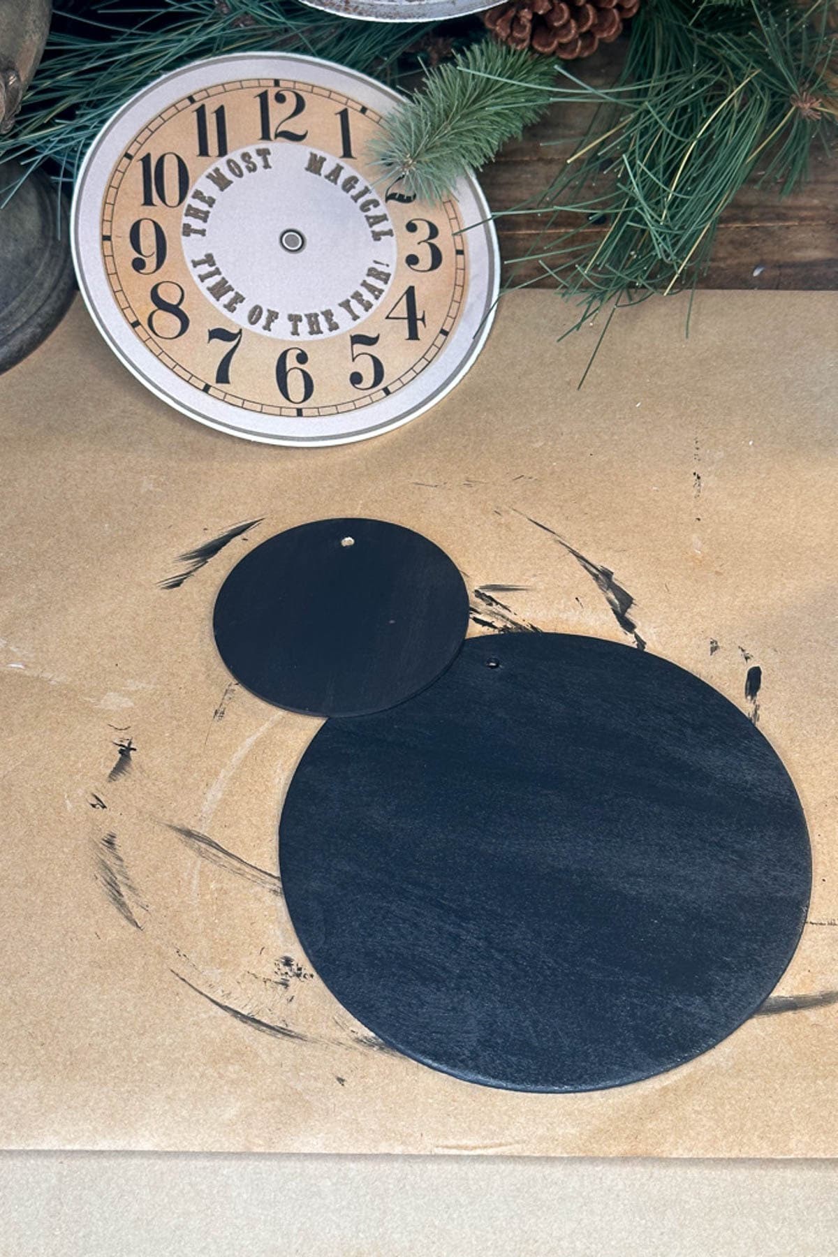 Two unfinished black clock faces with small holes in the center rest on a wooden surface, reminiscent of DIY projects. Black paint marks are visible around them, adding a vintage touch. In the background, part of a completed clock with large numbers and decorative greenery evokes a Christmas charm.