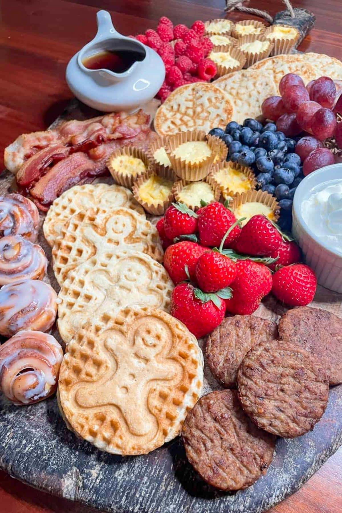 A breakfast board featuring waffles with gingerbread designs, sausage patties, bacon, mini cinnamon rolls, strawberries, blueberries, grapes, raspberries, mini muffins, syrup in a gravy boat, and a dish of whipped cream.