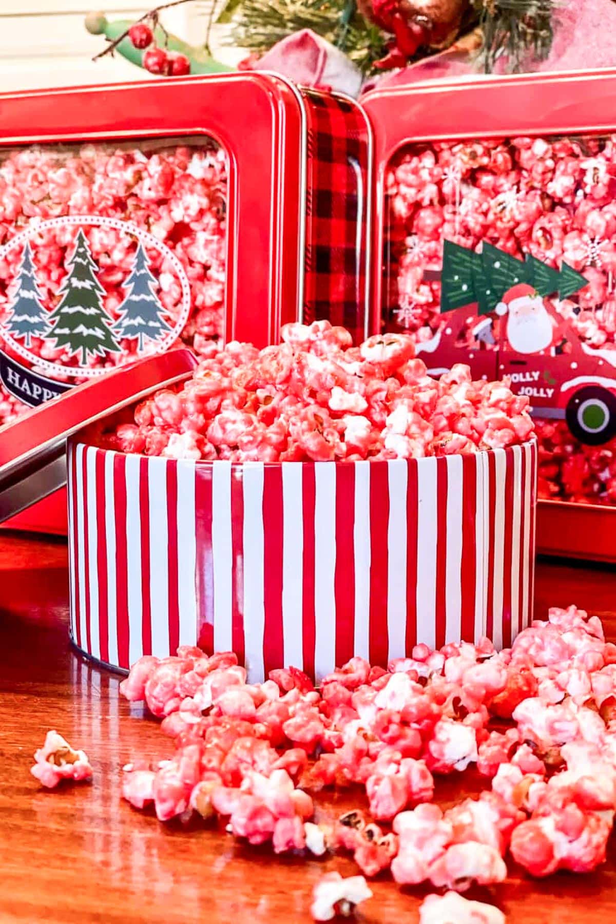 A red and white striped bowl filled with pink popcorn sits on a wooden surface. More popcorn is scattered around it. Behind the bowl are red tins decorated with festive holiday designs, including trees and a Santa motif.