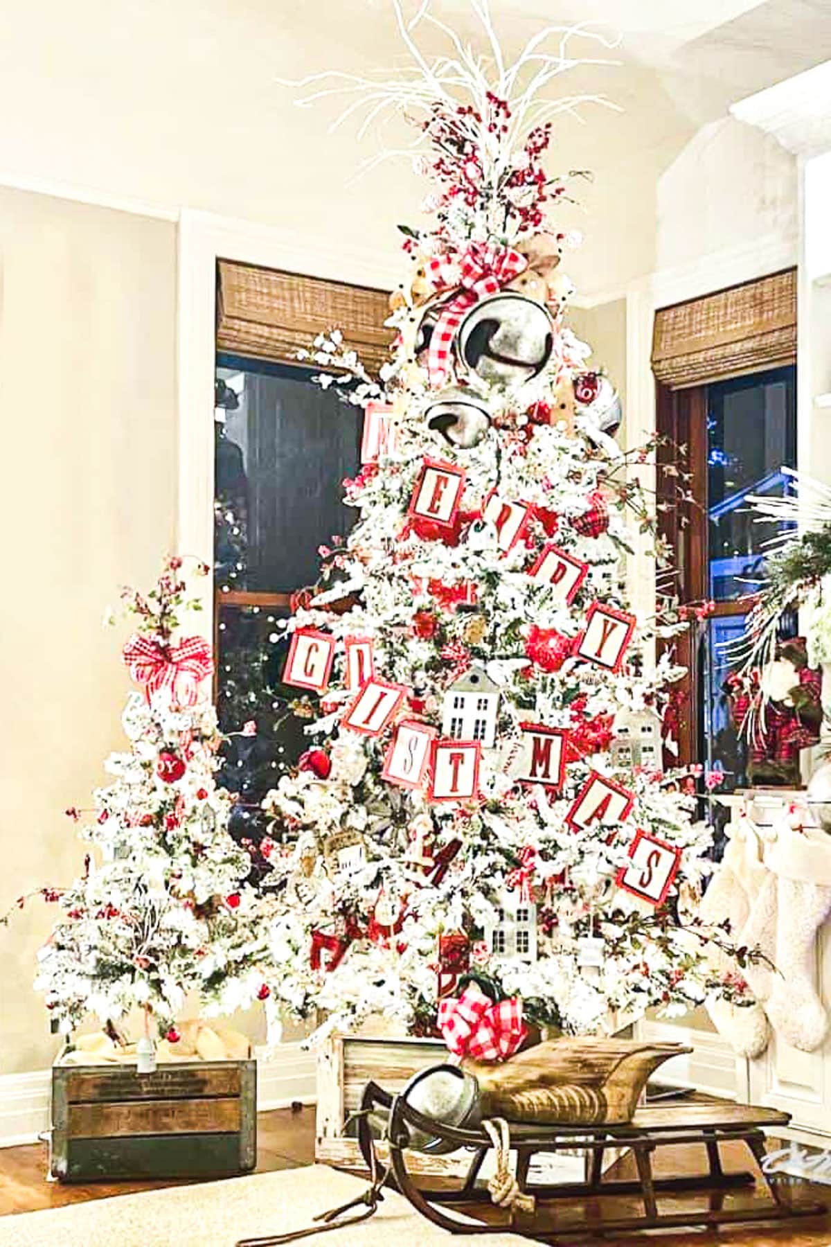 A Christmas scene with a large, elaborately decorated tree featuring red and white ornaments, a "Merry Christmas" sign, perfect for a grandkids' Christmas pajama party. A smaller matching tree nearby and a wooden sled in front complete the festive indoor display.