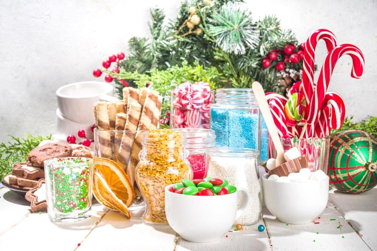 A festive display of holiday treats featuring candy canes, cookies, and jars filled with colorful sweets. The background is decorated with evergreen branches and red berries, creating a cheerful Christmas atmosphere.