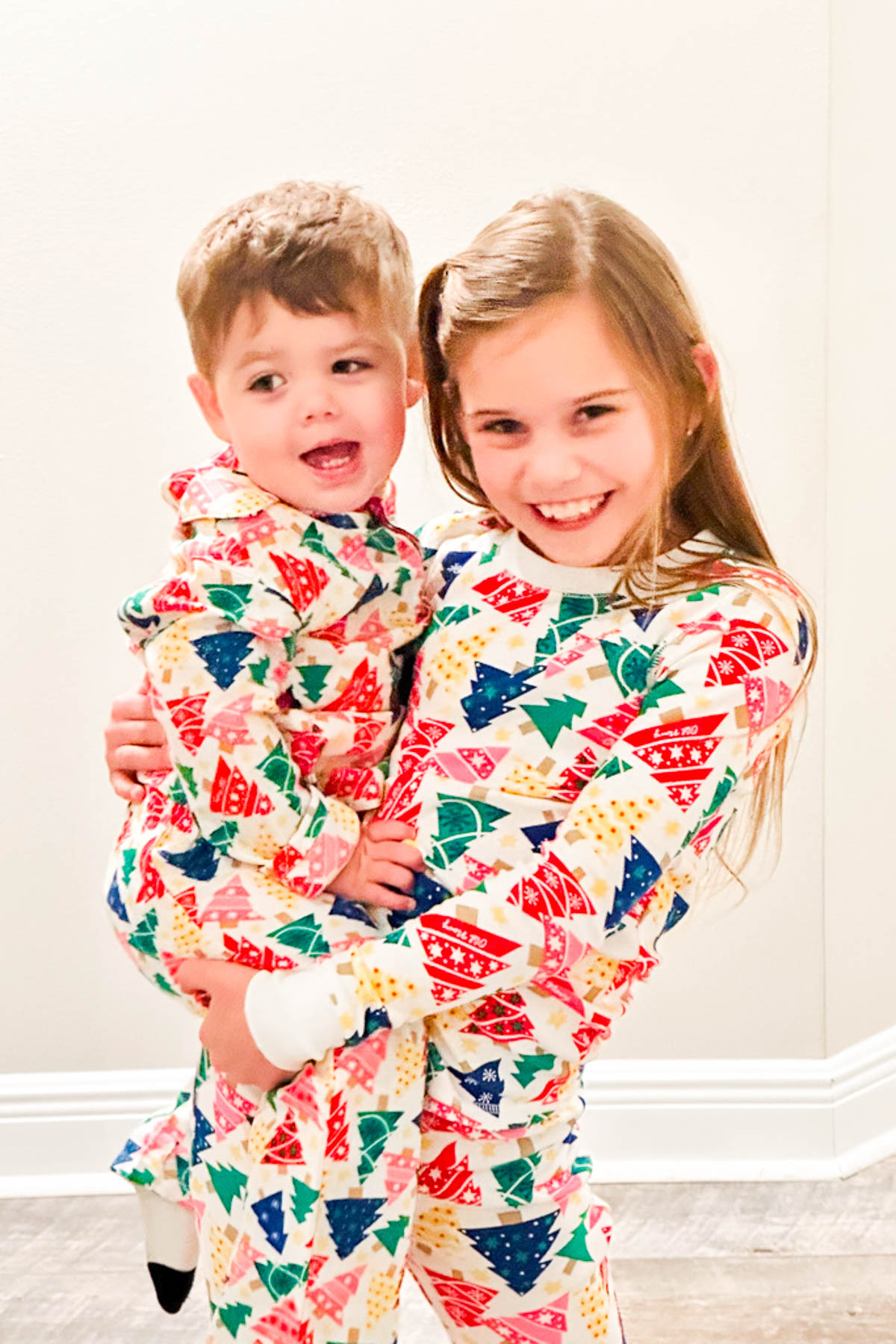 A young girl and a toddler, both wearing colorful, festive pajamas with Christmas tree patterns, smile in a warmly lit room during their Christmas Pajama Party. The girl is holding the toddler, and they both appear happy and cheerful.