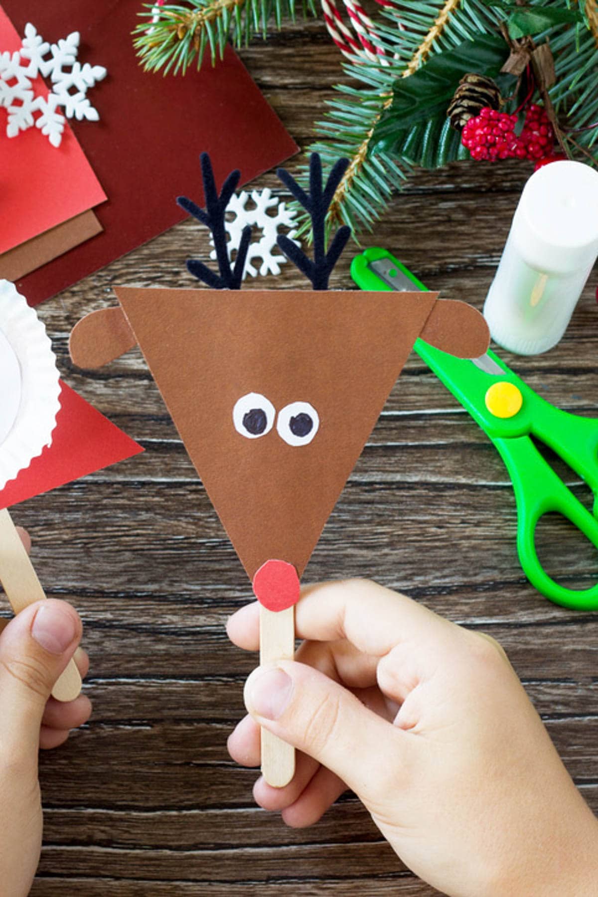 A person holds a handmade reindeer craft, fashioned from brown paper, a wooden stick, and red and black accents—an ideal project for grandkids. Scissors, glue, and festive decorations are scattered on the wooden table, sparking even more creative ideas for your Christmas pajama party.