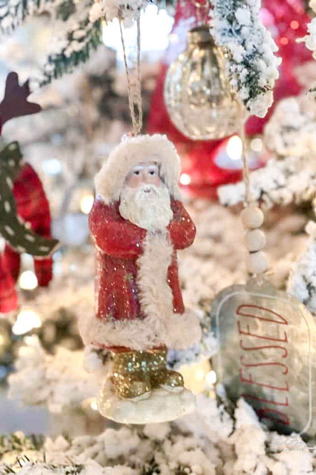 A close-up of a Christmas tree decorated with white snow, featuring a Santa Claus ornament in a red coat with white trim and glittery boots, perfectly matching the festive spirit of your Christmas Pajama Party. Nearby is a metal ornament embossed with "Blessed," alongside red and gold baubles.